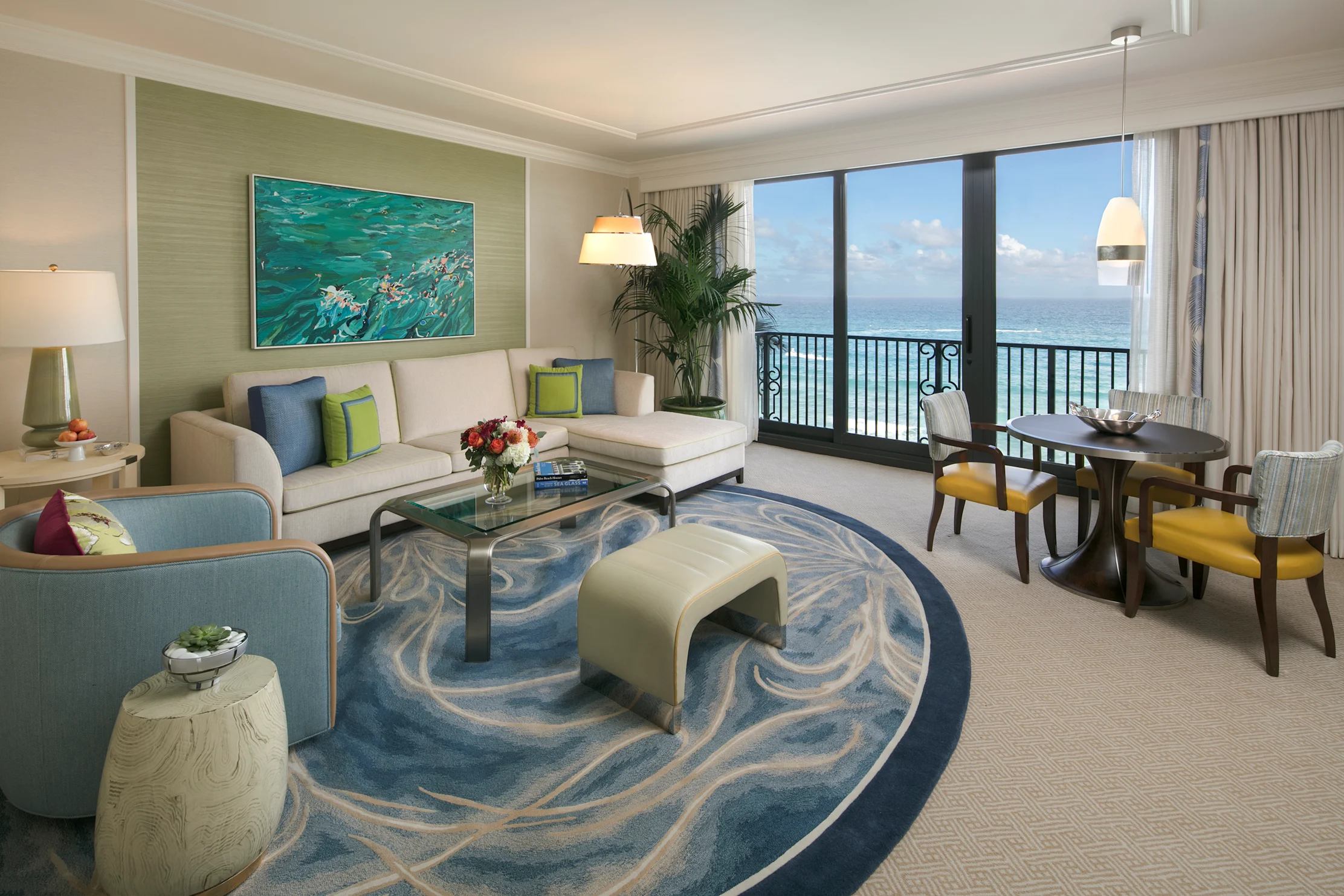 Modern hotel suite with ocean view, beige sofa, blue wave rug, dining table, and coastal artwork.