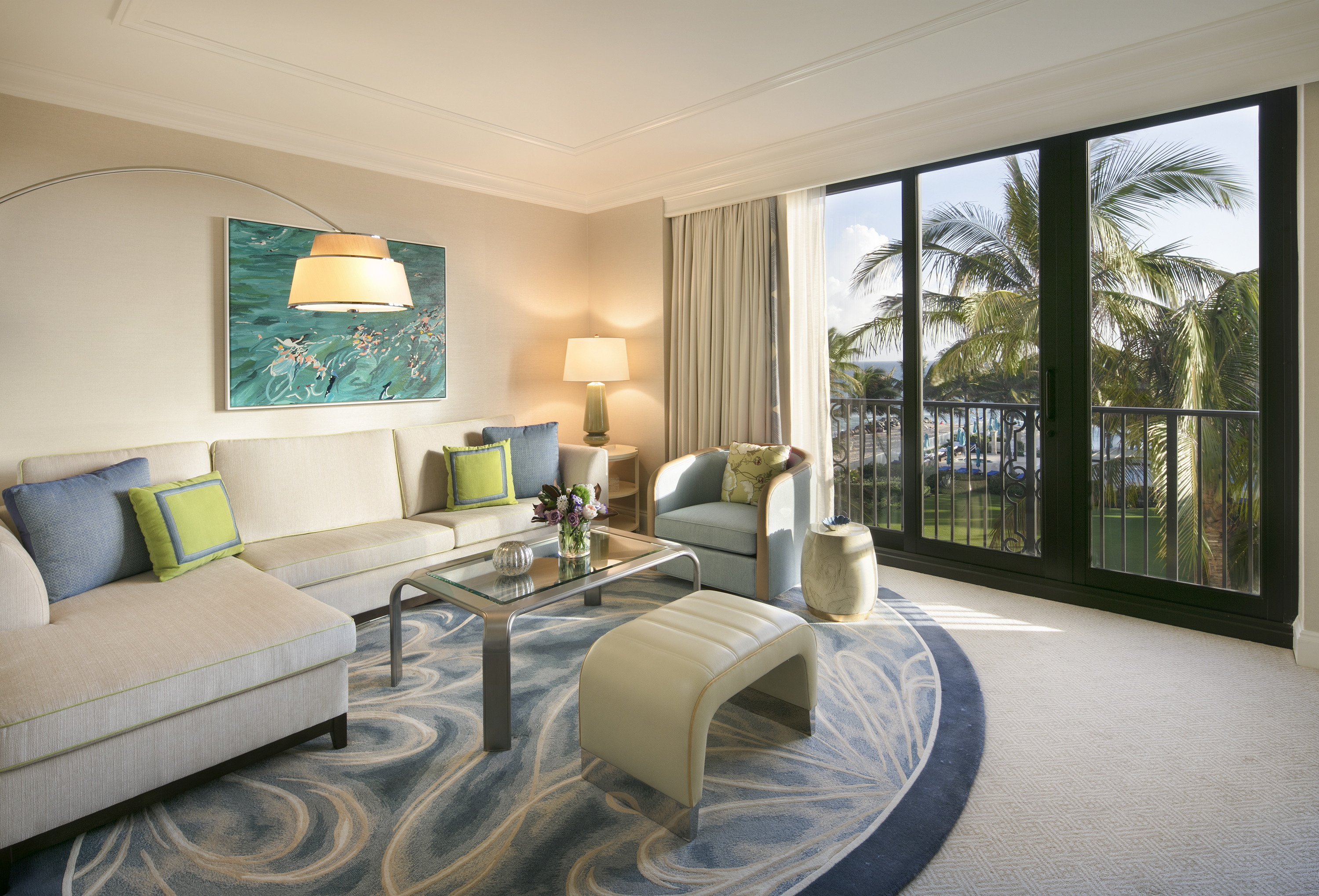 Bright hotel living room with beige sofa, blue and green pillows, ocean artwork, and balcony view of palm trees.