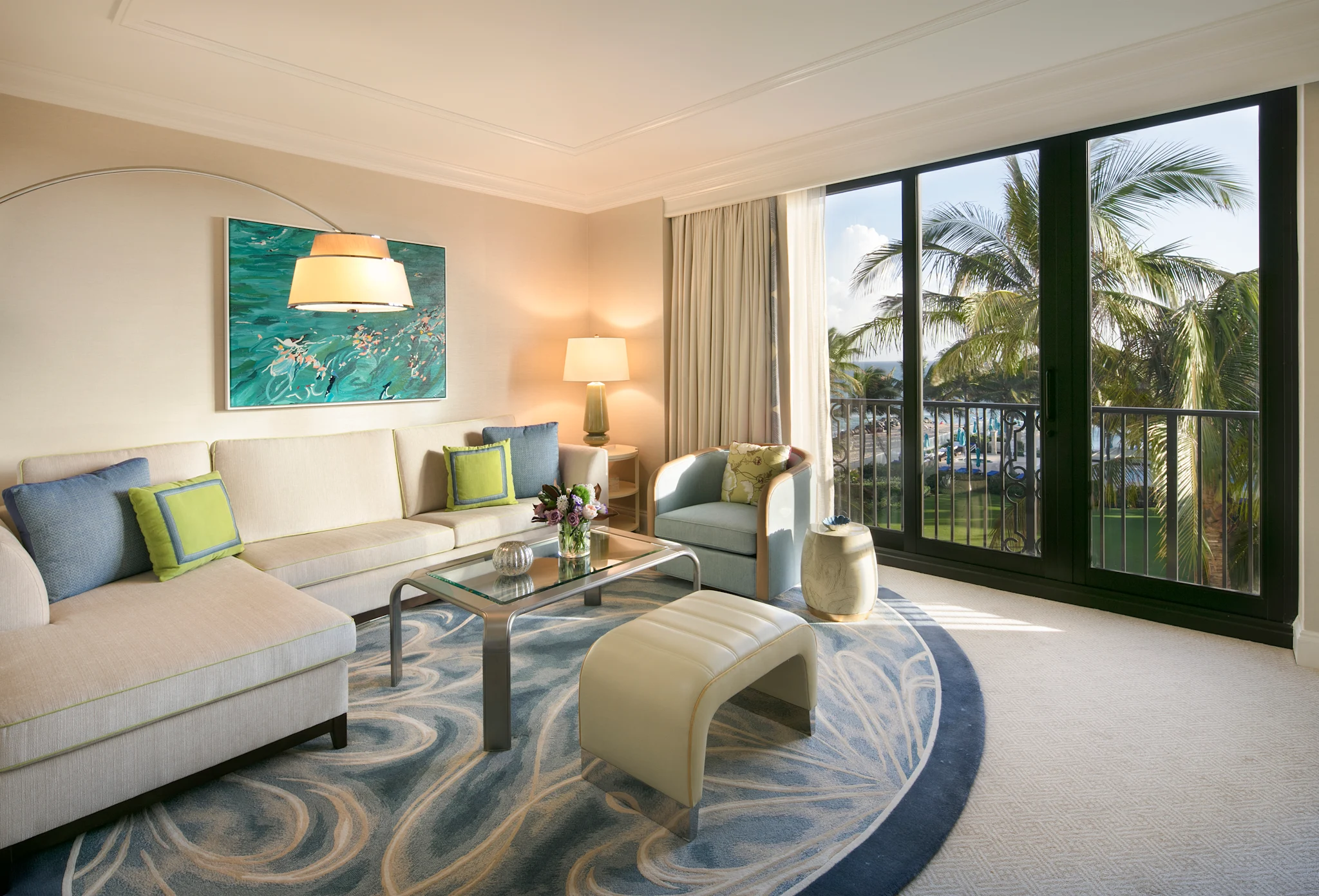 Bright hotel living room with beige sofa, blue and green pillows, ocean artwork, and balcony view of palm trees.