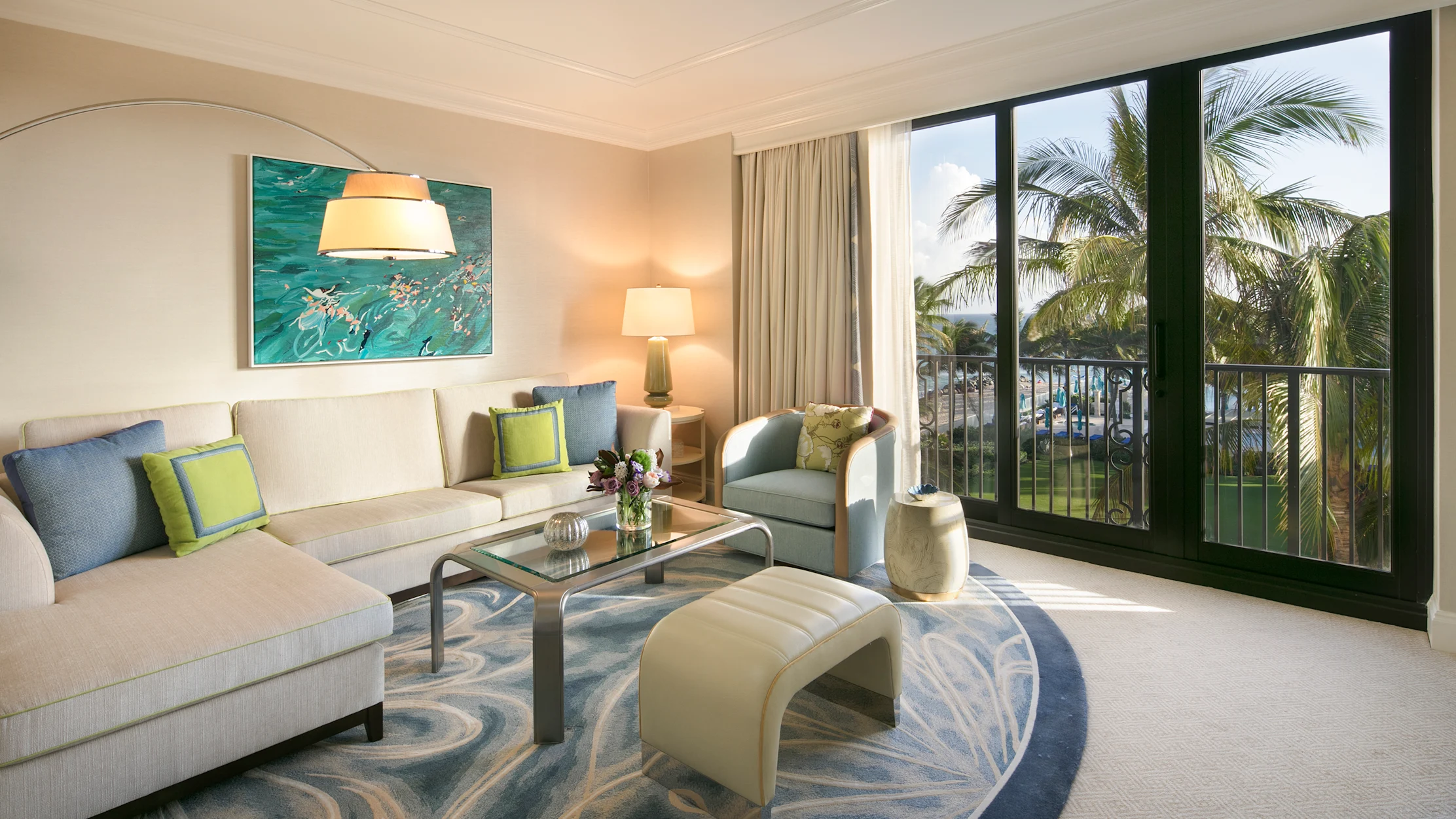 Bright hotel living room with beige sofa, blue and green pillows, ocean artwork, and balcony view of palm trees.