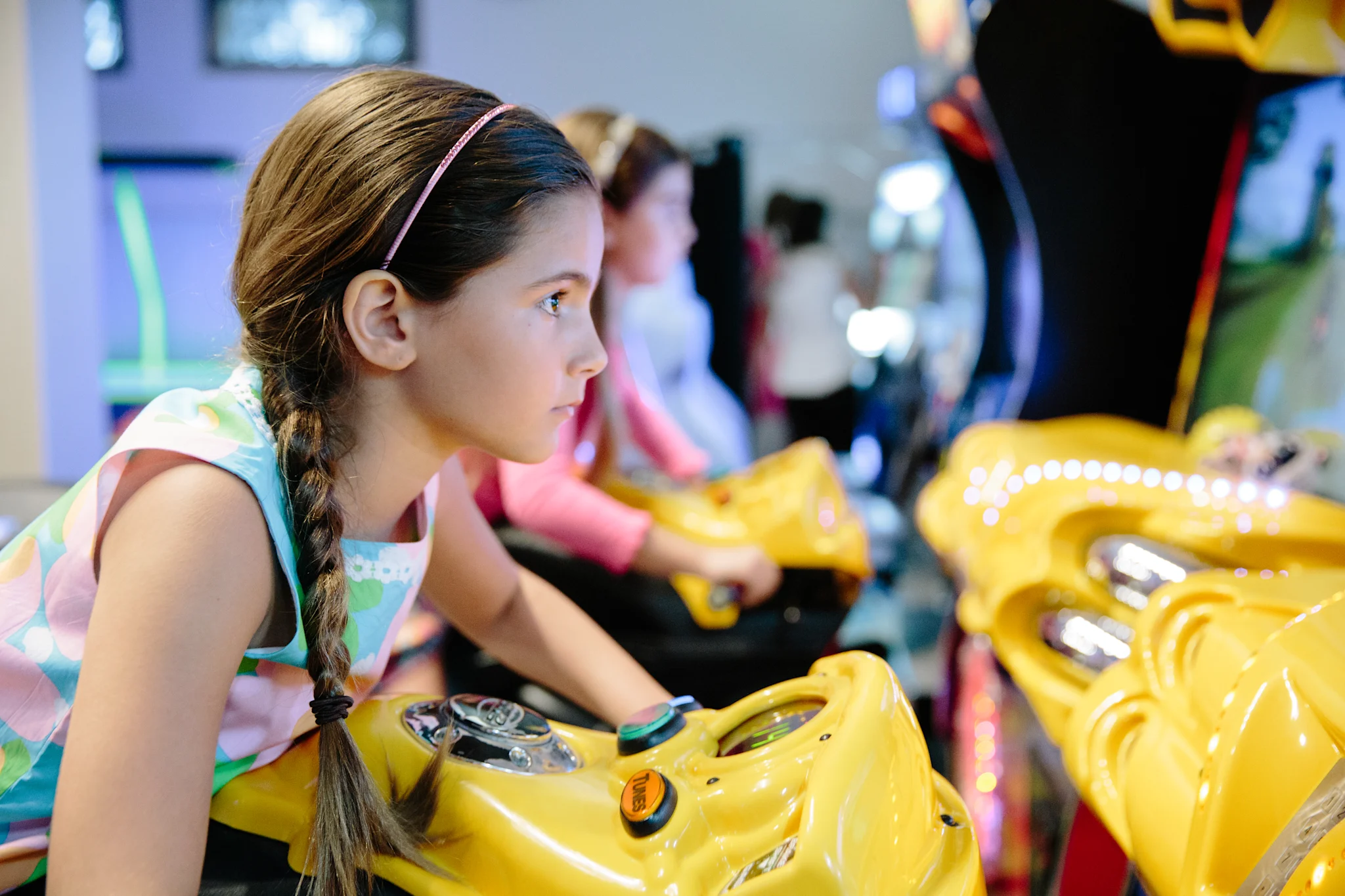 Young girl with braided hair playing yellow arcade racing game in entertainment center.