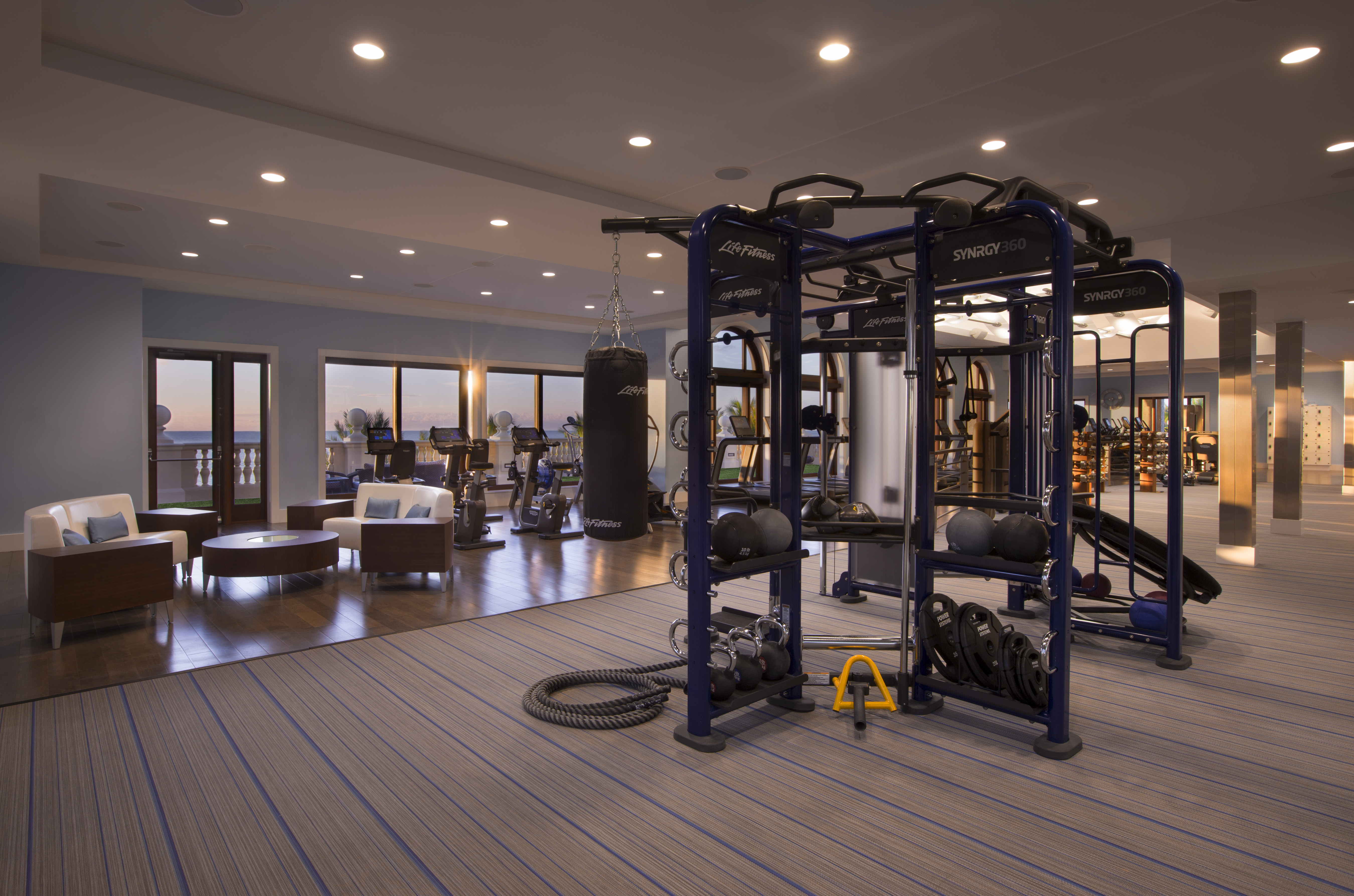 Modern fitness center with blue exercise equipment, lounge seating, and ocean view through glass doors.