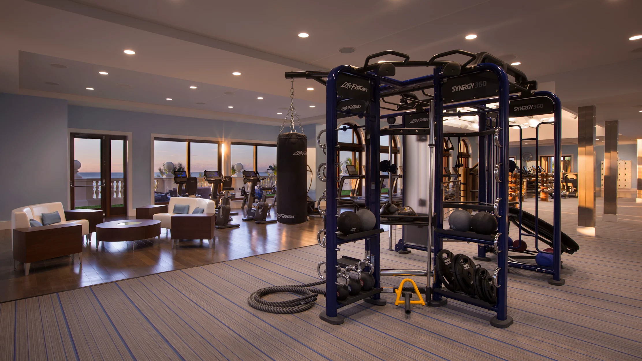 Modern fitness center with blue exercise equipment, lounge seating, and ocean view through glass doors.