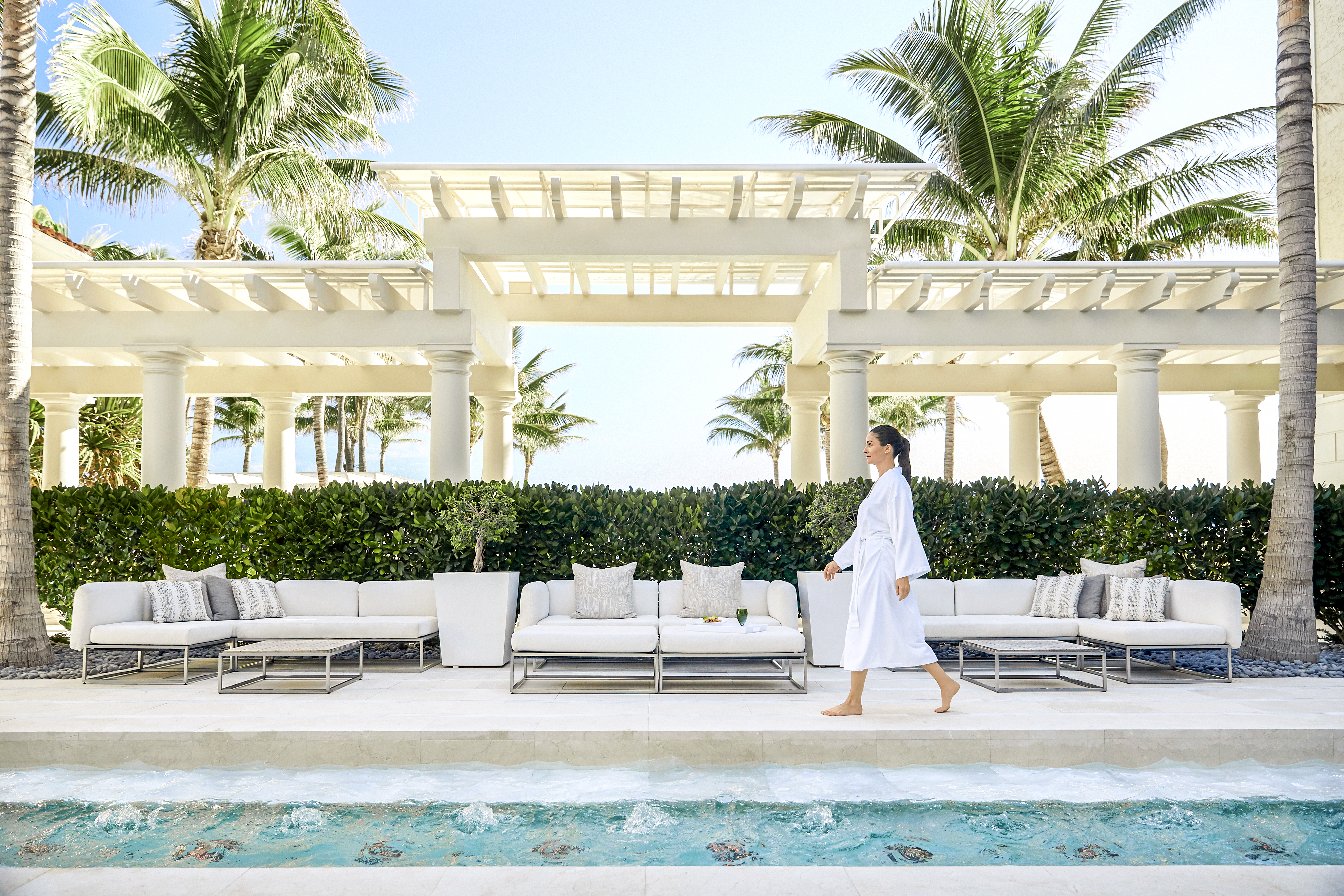 Woman in white robe walking by luxury pool with white outdoor furniture and palm trees at resort.