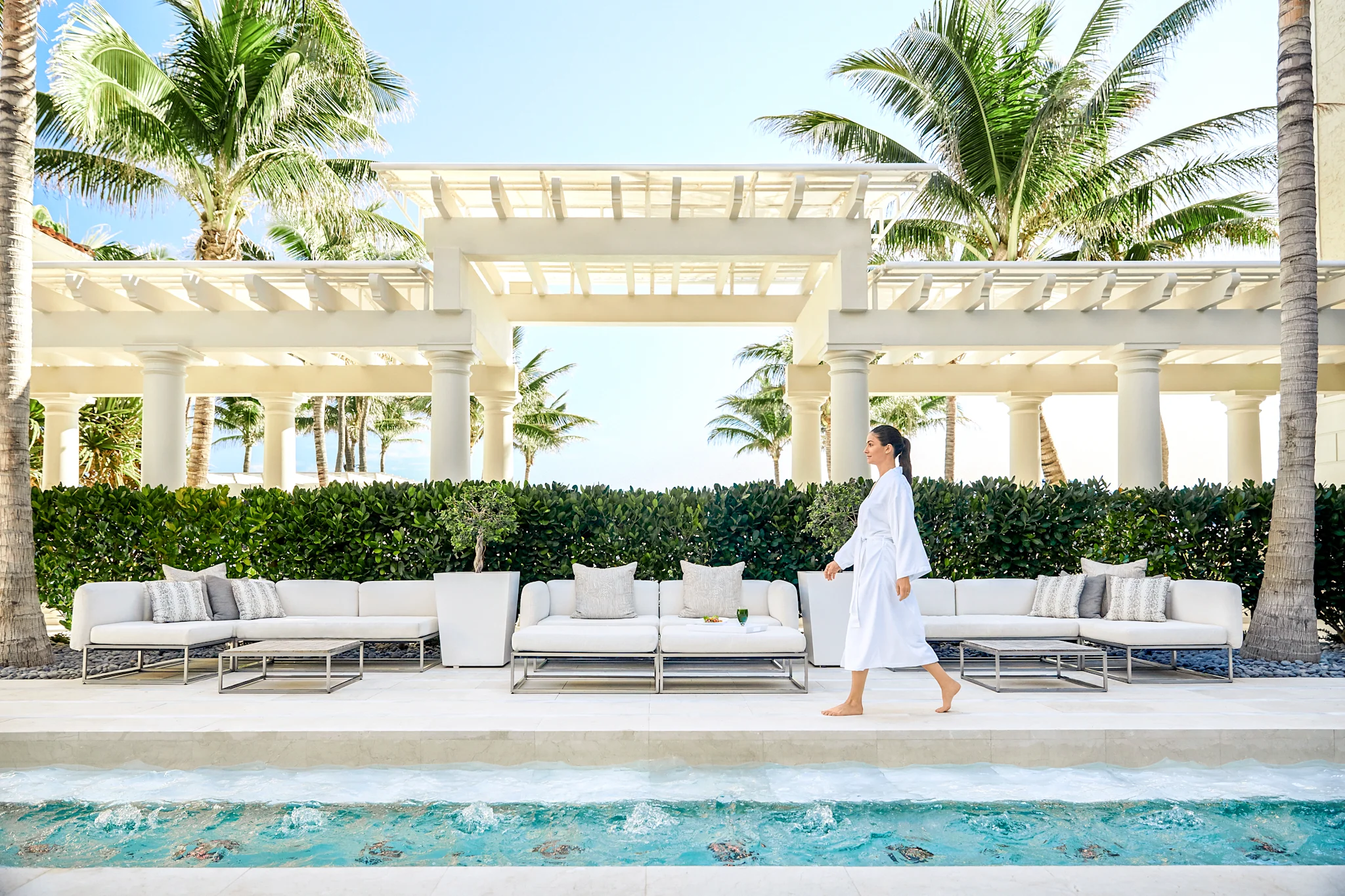 Woman in white robe walking by luxury pool with white outdoor furniture and palm trees at resort.