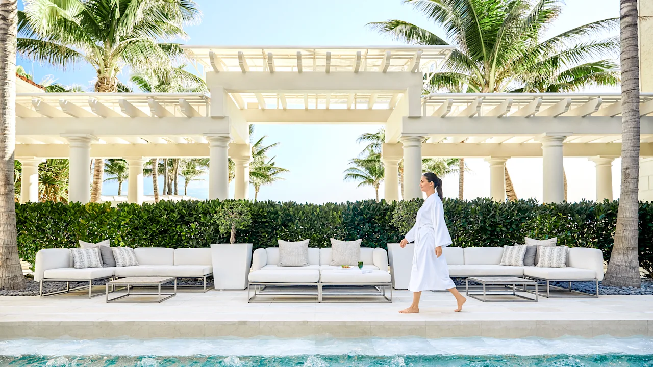 Woman in white robe walking by luxury pool with white outdoor furniture and palm trees at resort.