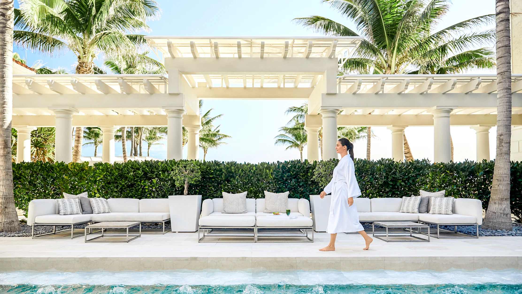 Woman in white robe walking by luxury pool with white outdoor furniture and palm trees at resort.