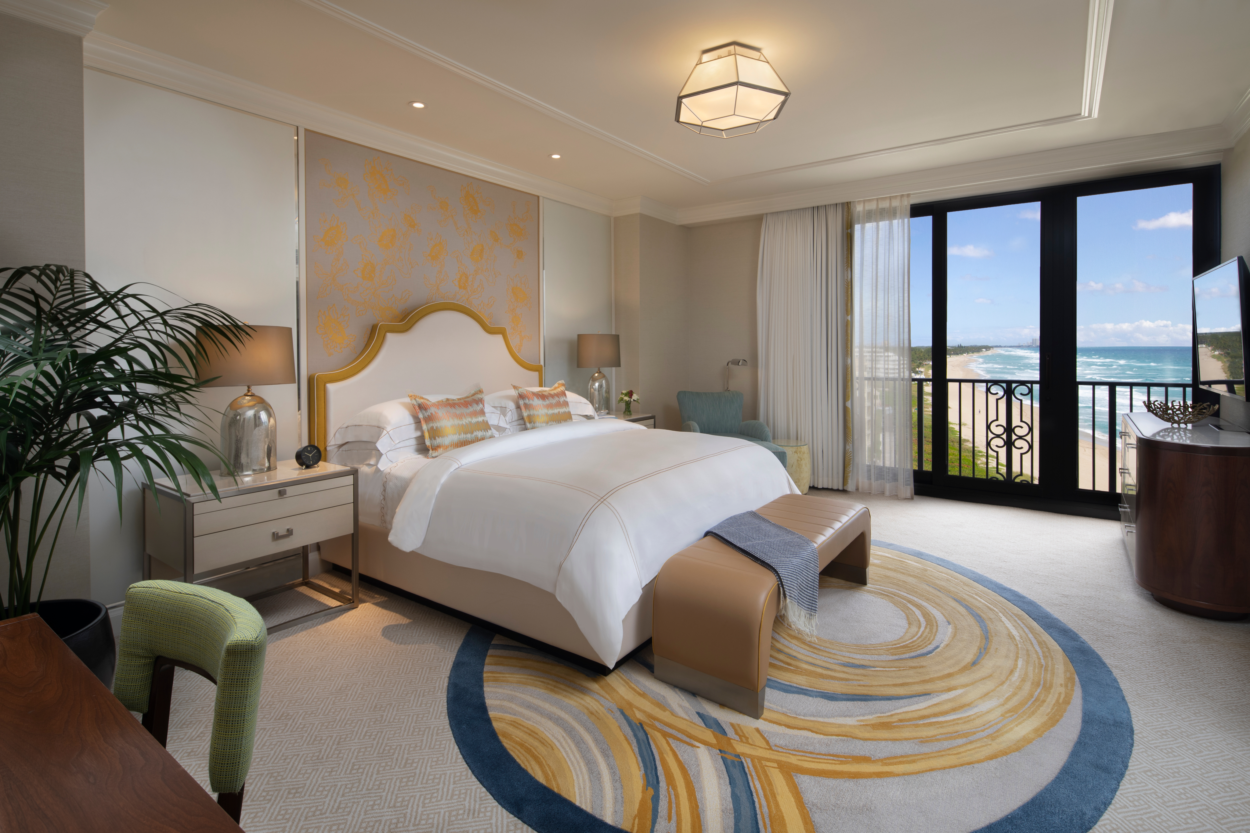 Luxury hotel bedroom with ocean view balcony, white bed with gold headboard, and decorative rug.