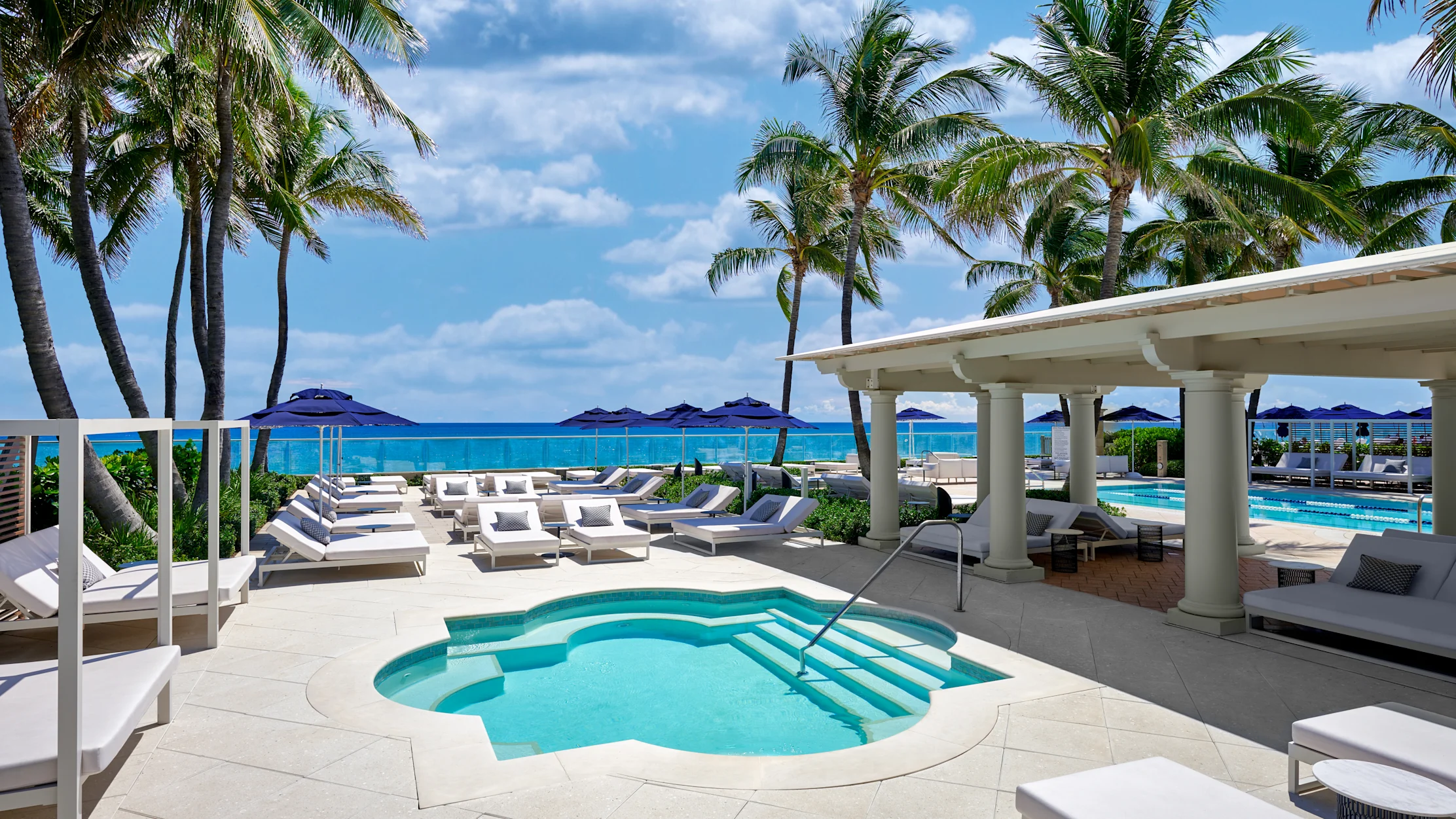 Luxury beachfront resort with turquoise pool, white loungers, palm trees, and ocean view under blue sky.