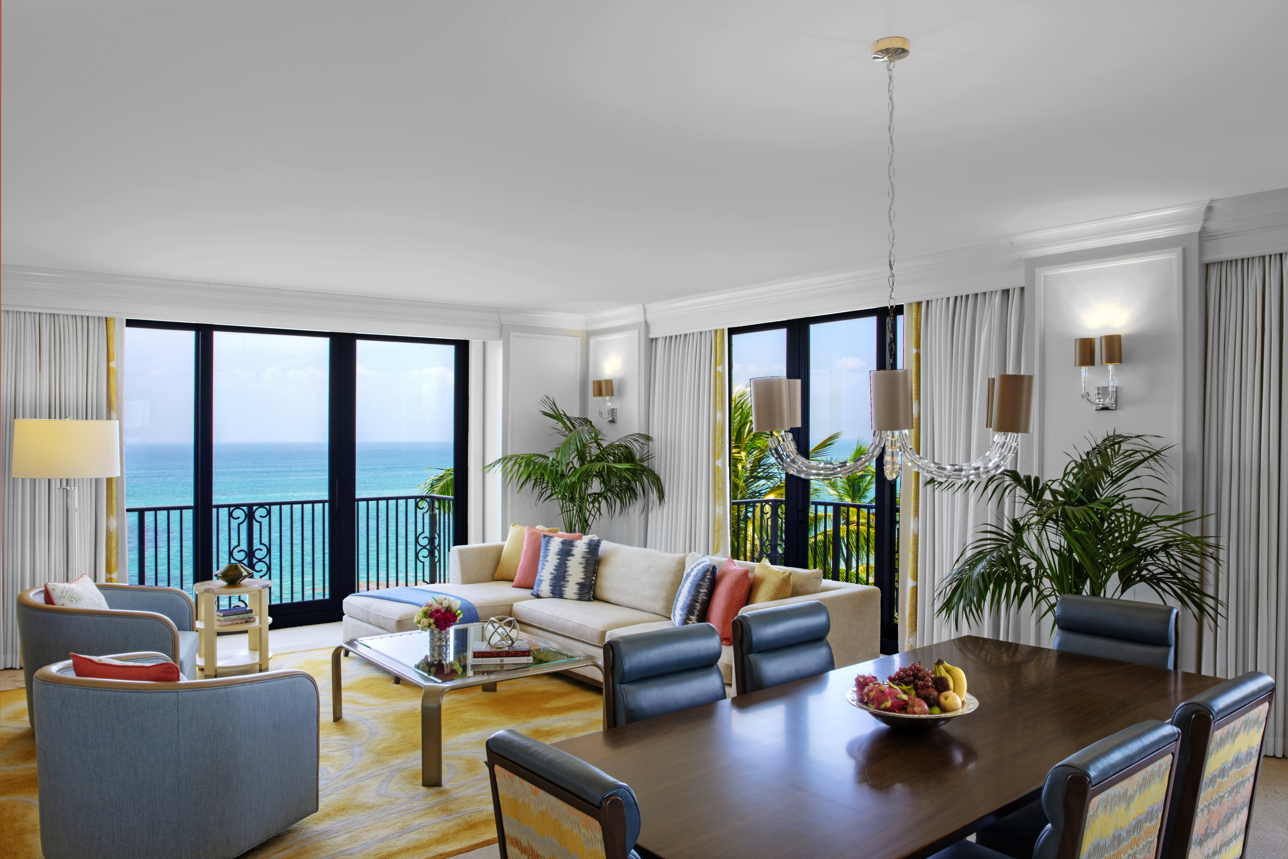 Bright hotel suite with ocean view, white sofa, dining table, and tropical plants by windows.