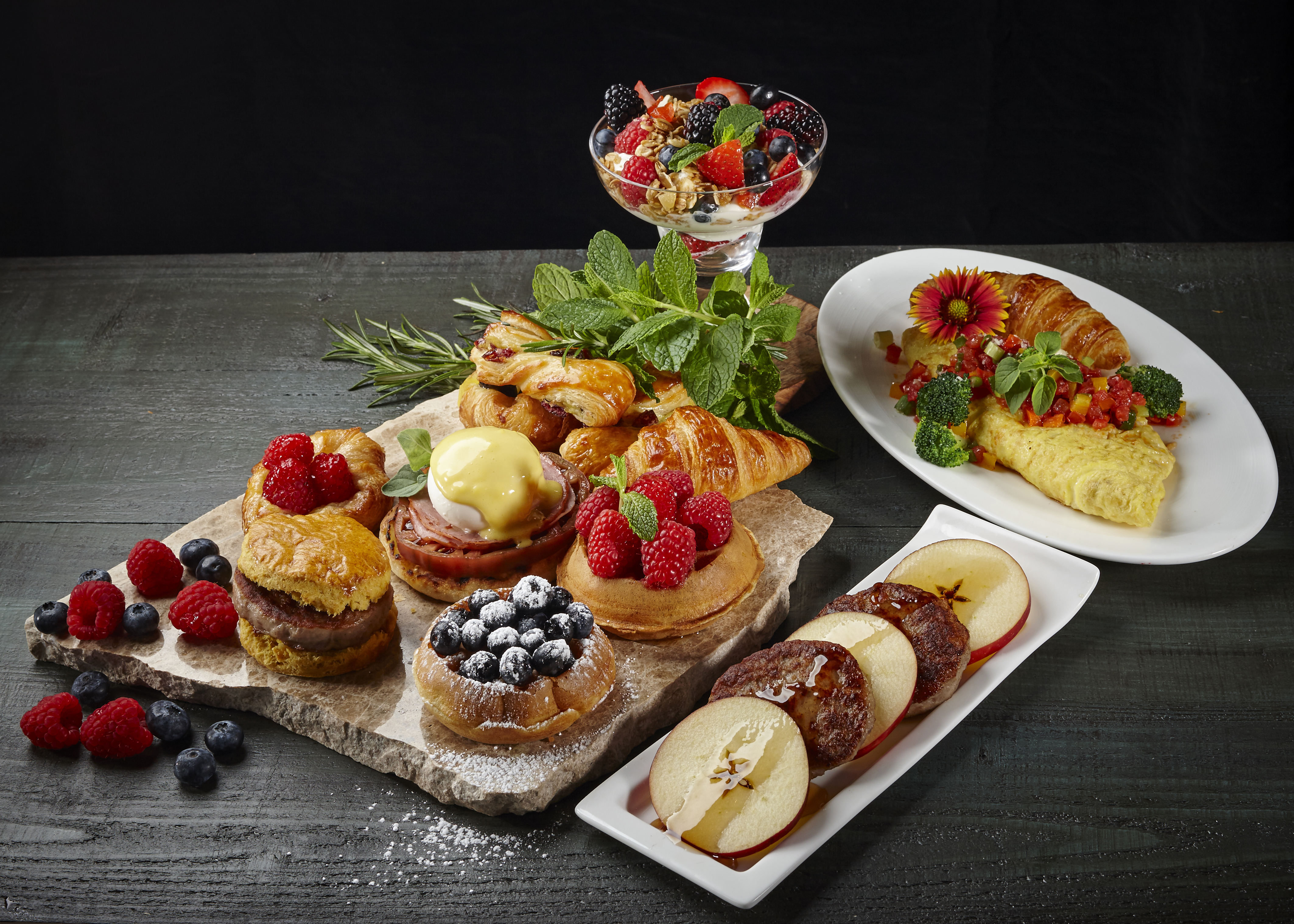 Gourmet breakfast spread with pastries, berries, omelet, and fruit parfait on dark wood table.