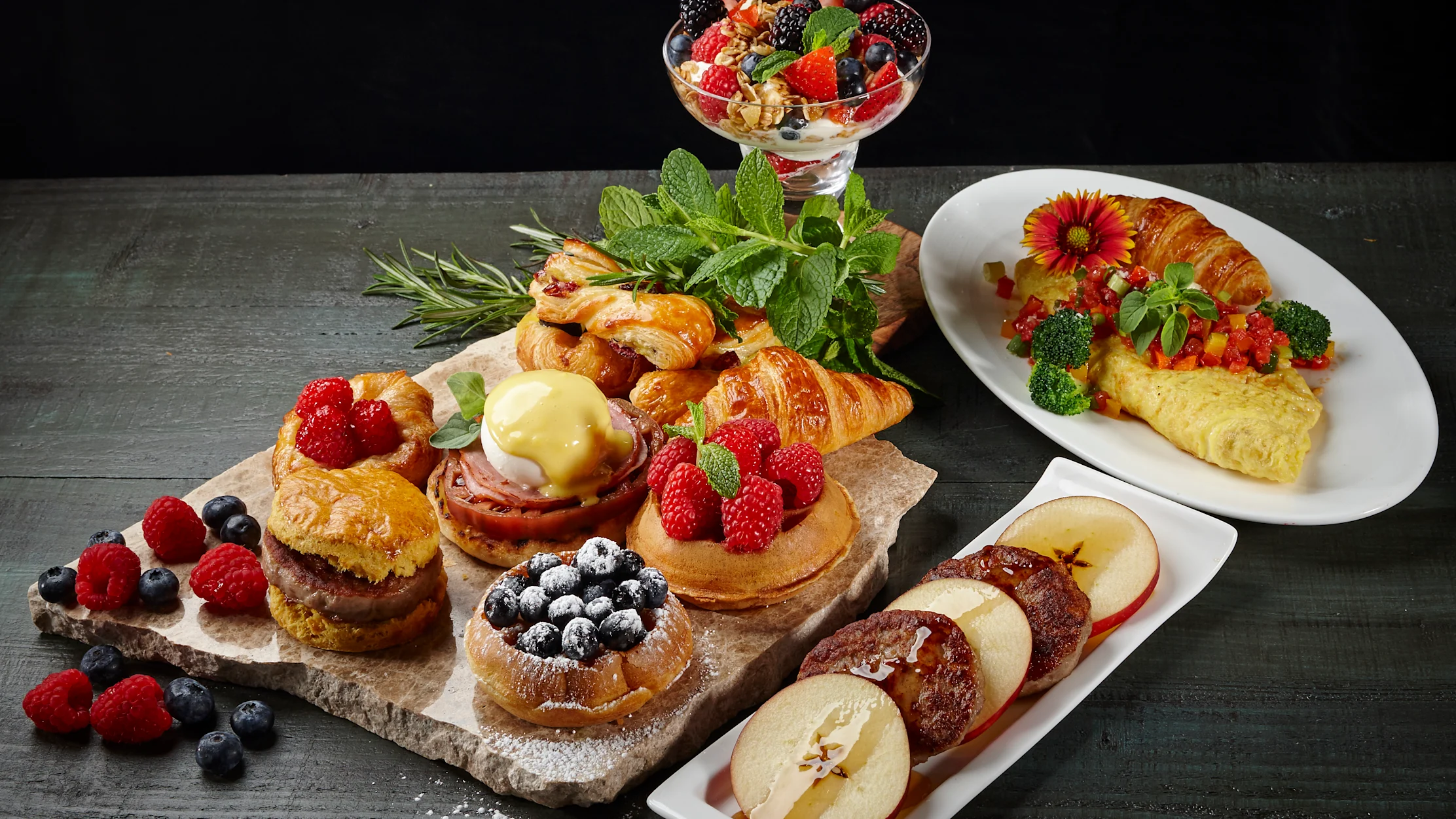 Gourmet breakfast spread with pastries, berries, omelet, and fruit parfait on dark wood table.