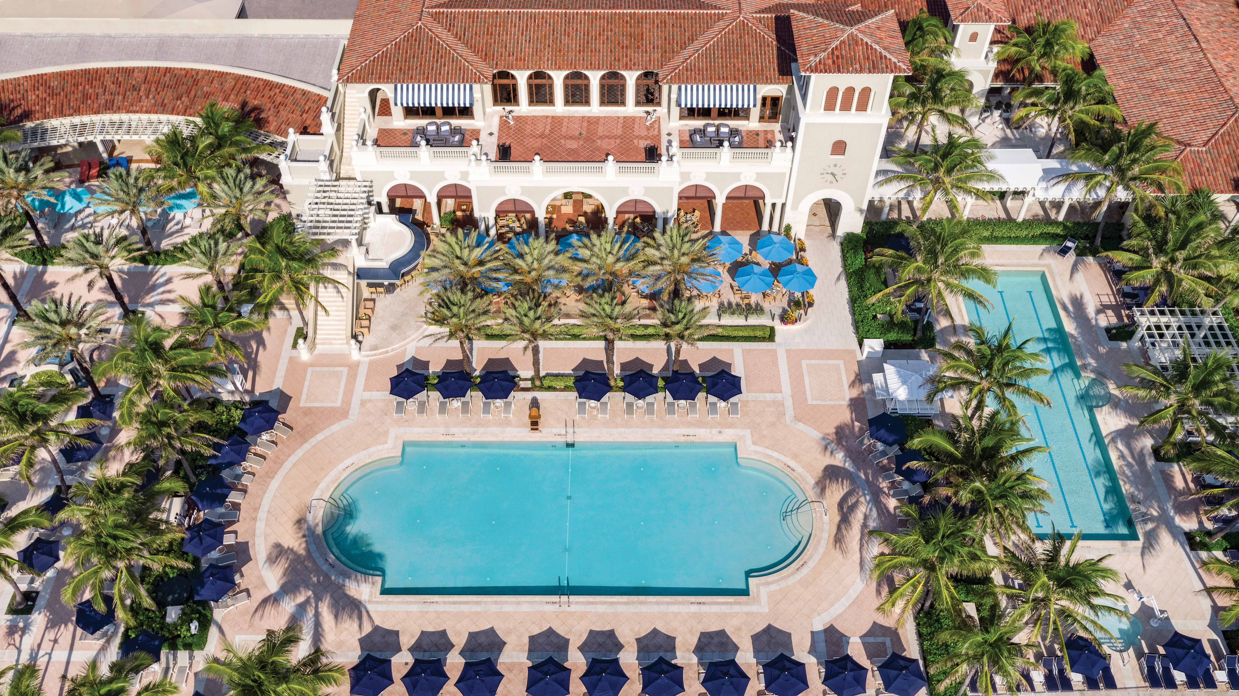 Aerial view of a Mediterranean-style resort with white buildings, red tile roofs, blue pool, and palm trees.