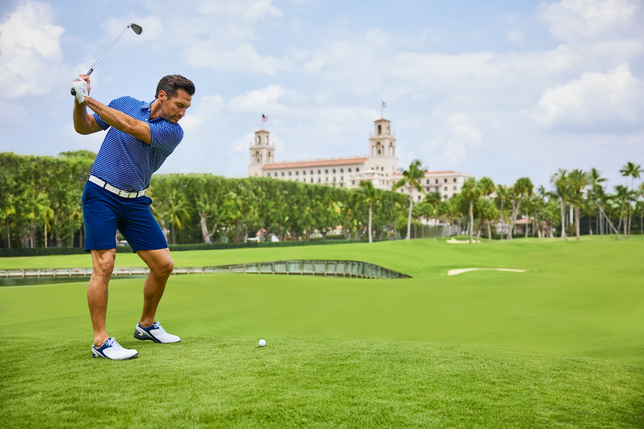 Golfer in blue striped shirt mid-swing on green course with historic building and palm trees.