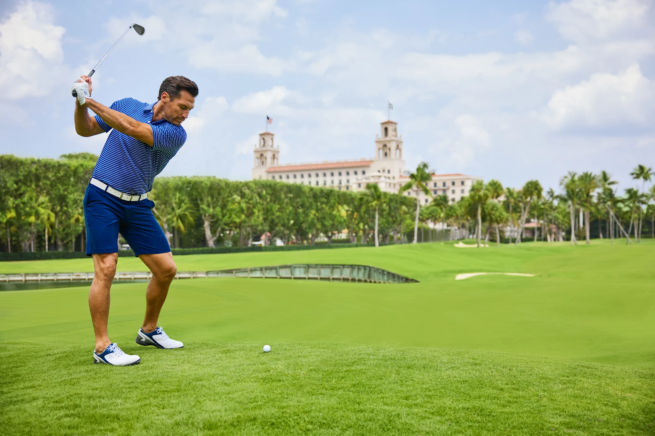 Golfer in blue striped shirt mid-swing on green course with historic building and palm trees.