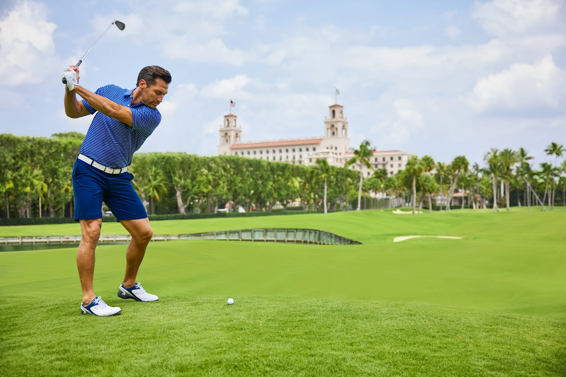 Golfer in blue striped shirt mid-swing on green course with historic building and palm trees.