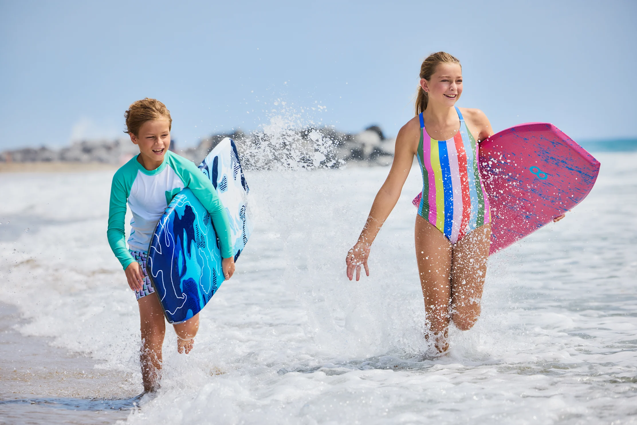 Two children running through shallow beach water carrying colorful boogie boards and smiling.