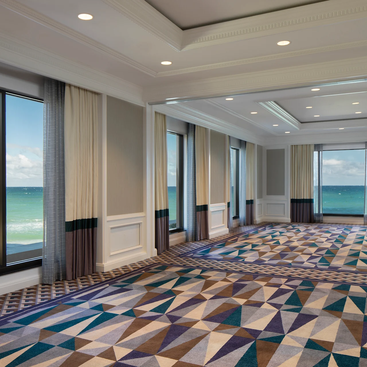 Elegant ballroom with geometric patterned carpet, floor-to-ceiling windows, ocean views, and recessed lighting.