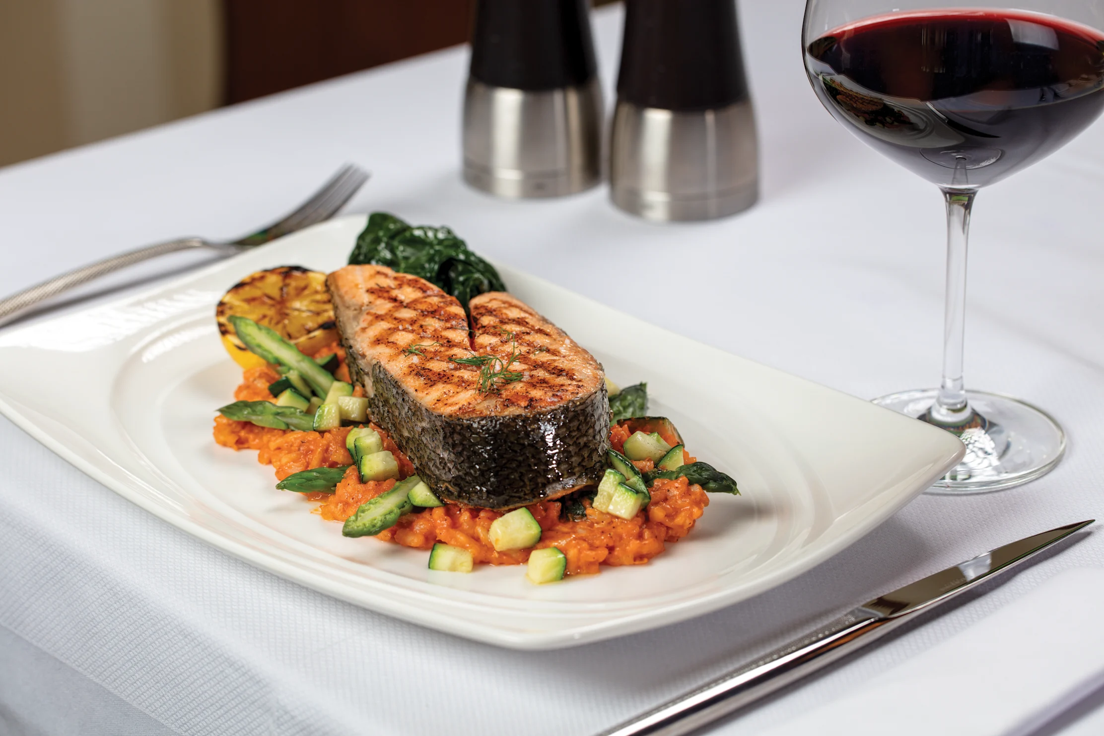 Grilled salmon steak on orange risotto with vegetables and a glass of red wine on white plate.