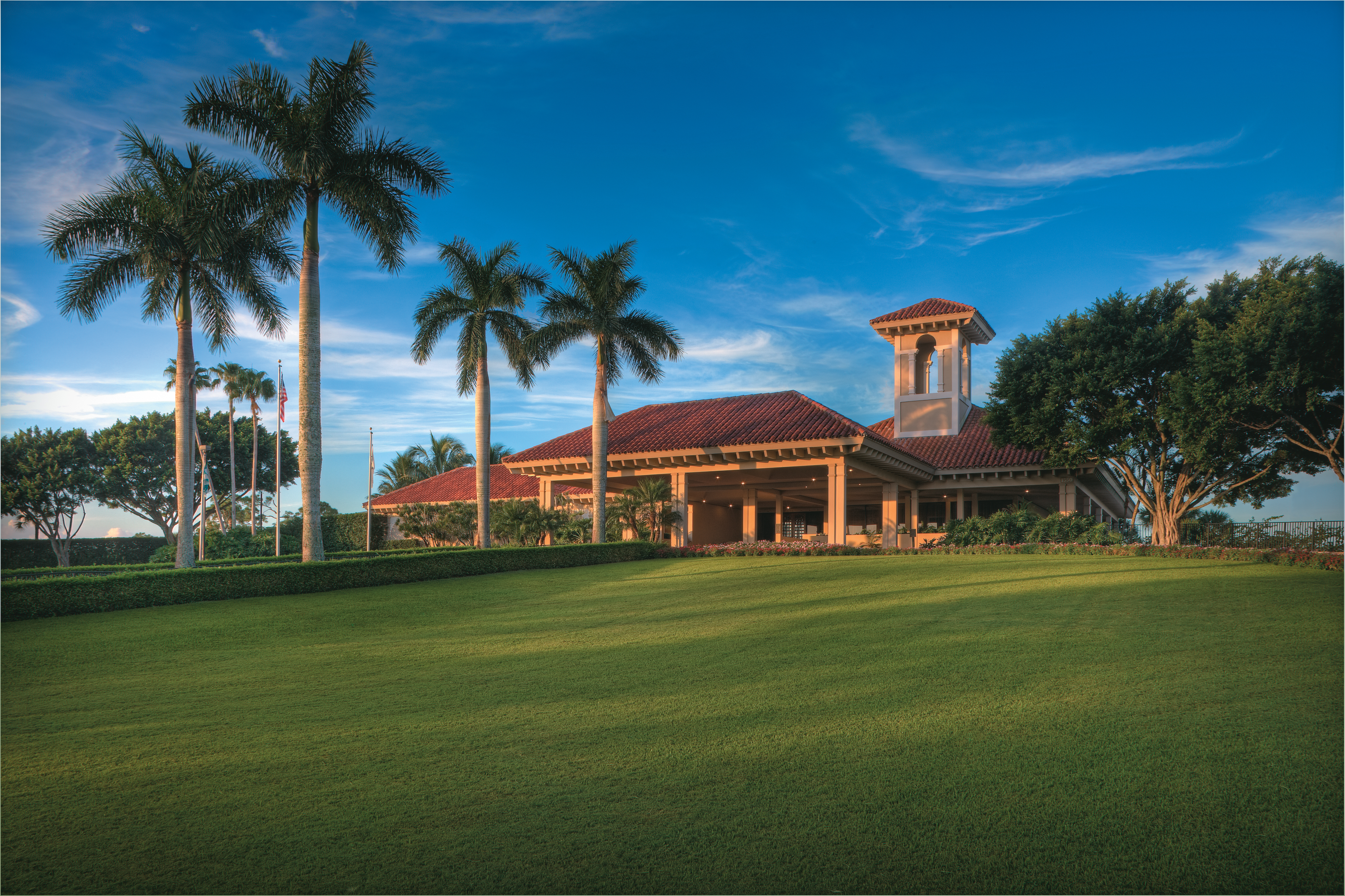 Mediterranean-style clubhouse with red tile roof and bell tower, surrounded by palm trees and green lawn.
