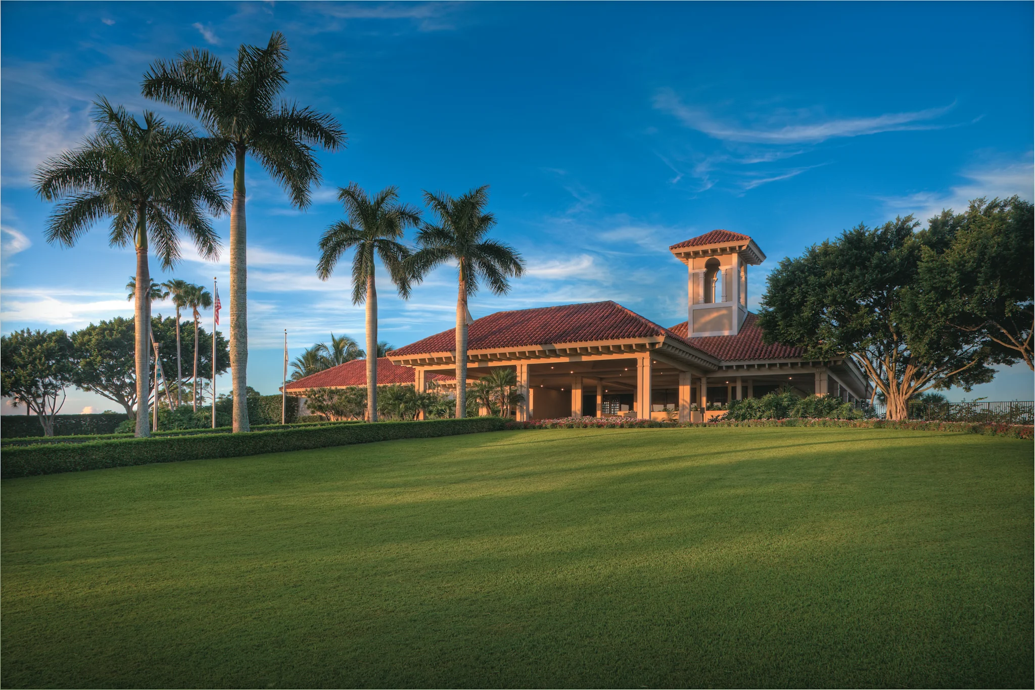 Mediterranean-style clubhouse with red tile roof and bell tower, surrounded by palm trees and green lawn.