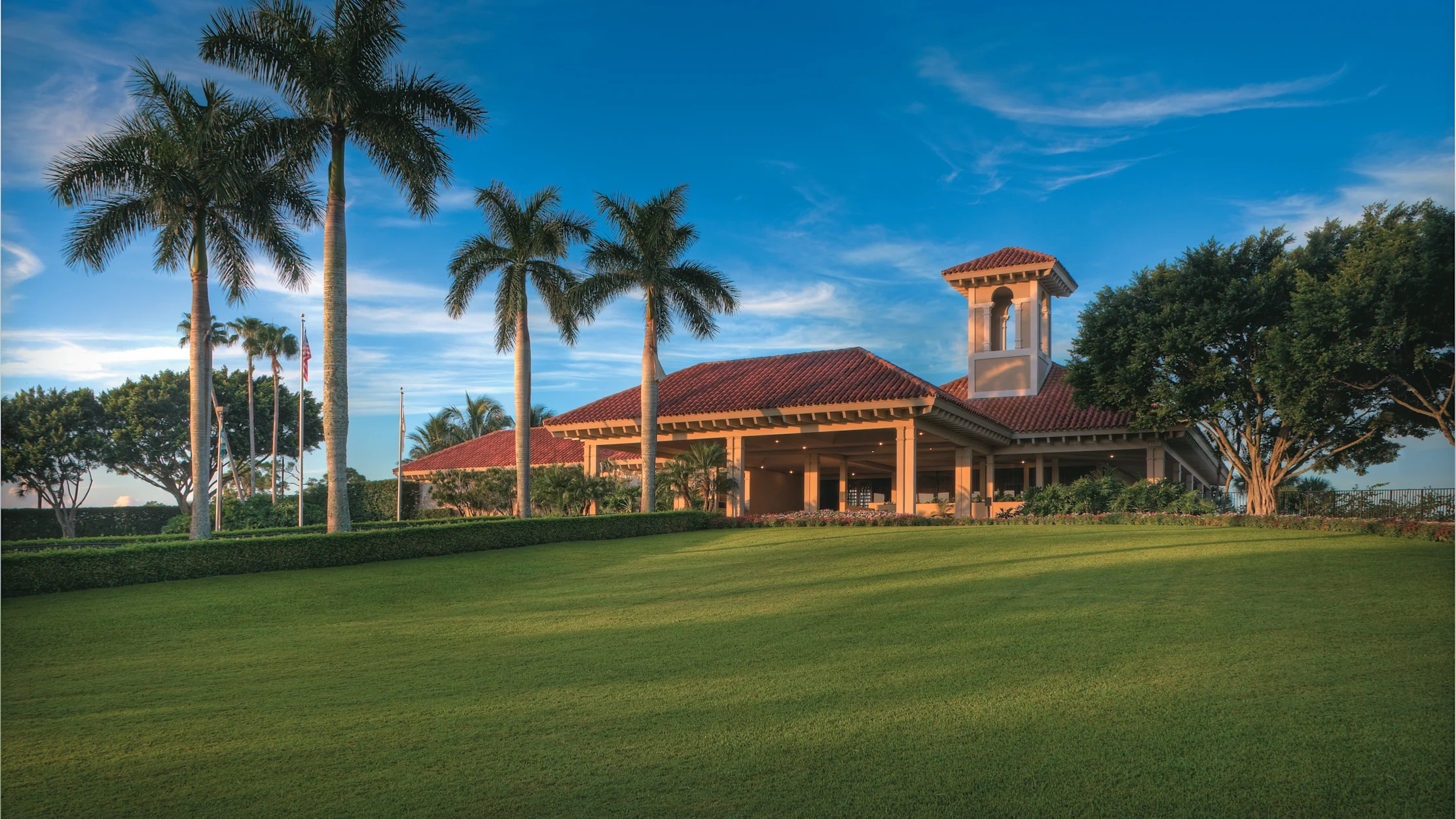 Mediterranean-style clubhouse with red tile roof and bell tower, surrounded by palm trees and green lawn.