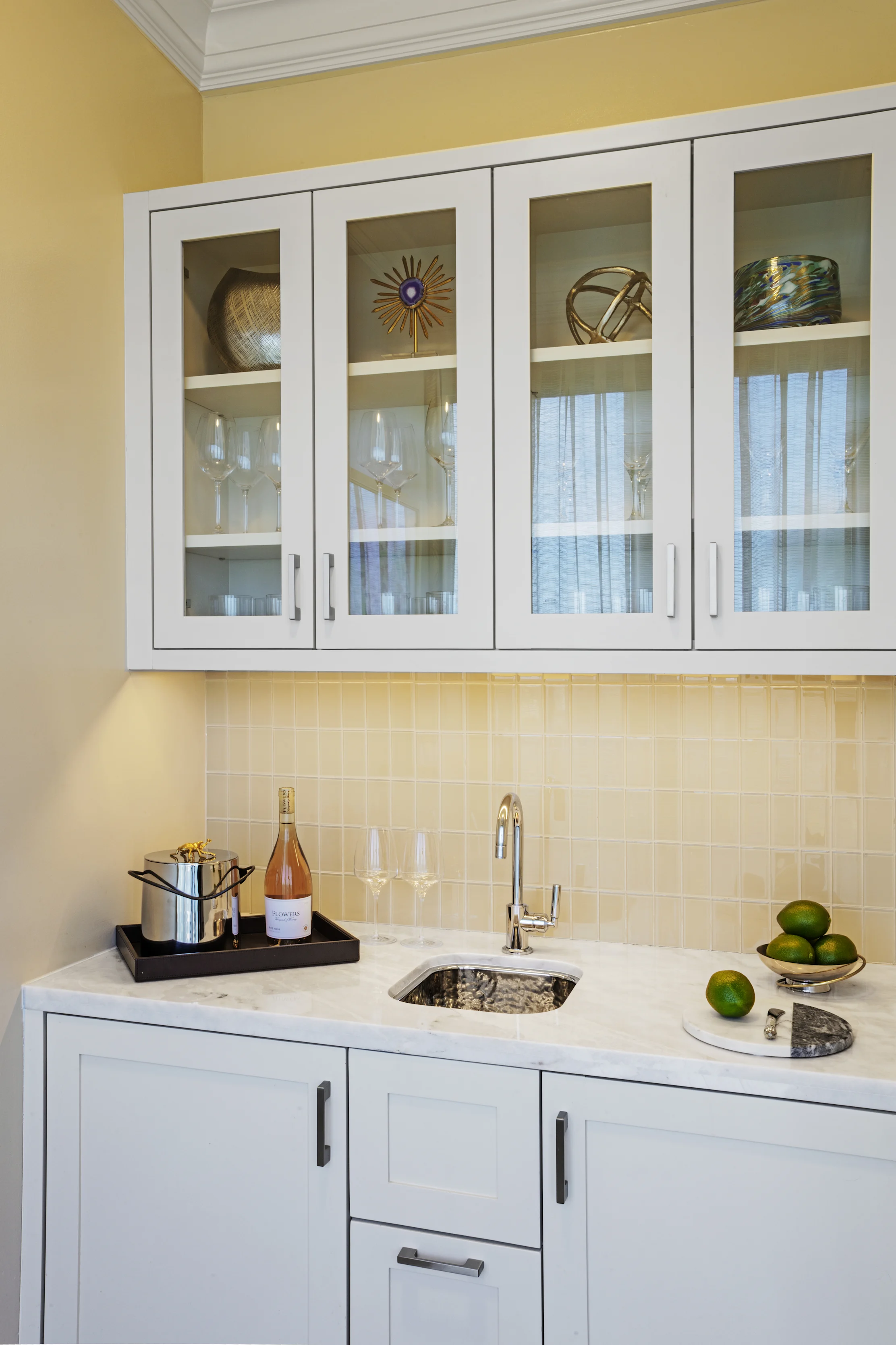 White kitchen bar area with glass-front cabinets, marble countertop, sink, wine bottle, and limes.