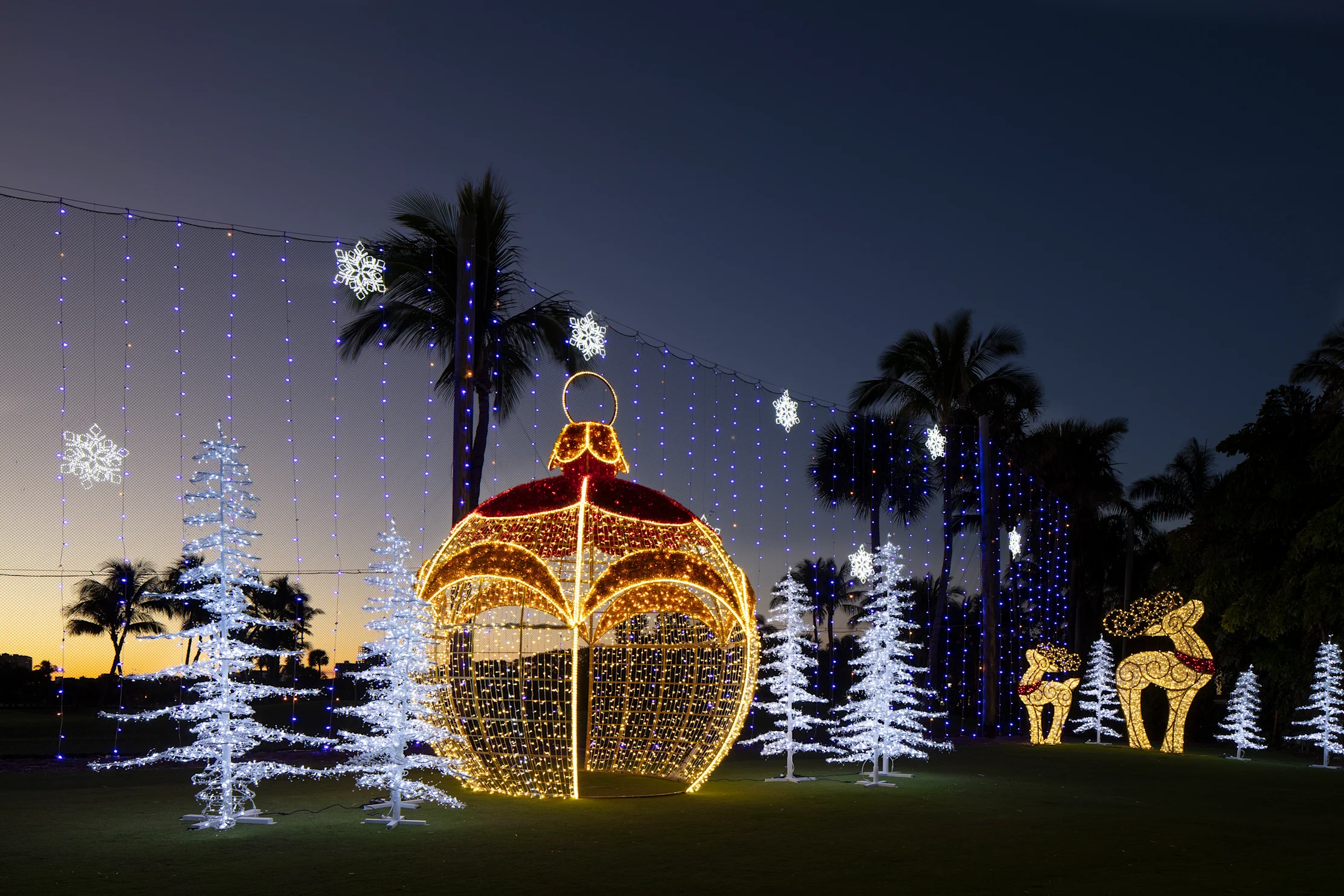 Giant illuminated Christmas ornament with white trees, snowflakes, and reindeer lights at dusk.