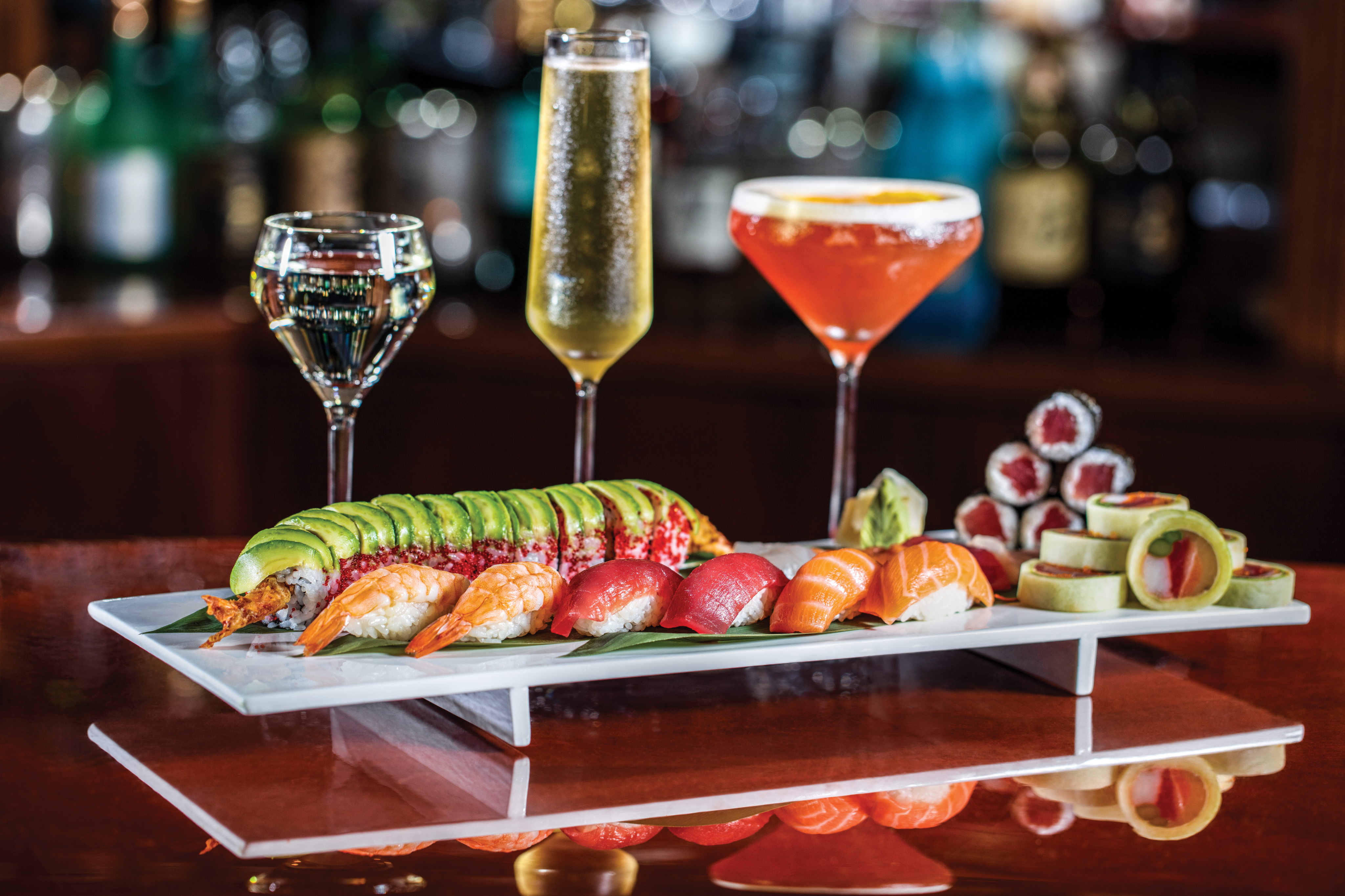 Assorted sushi platter with three colorful cocktails on a bar counter with blurred bottles in background.
