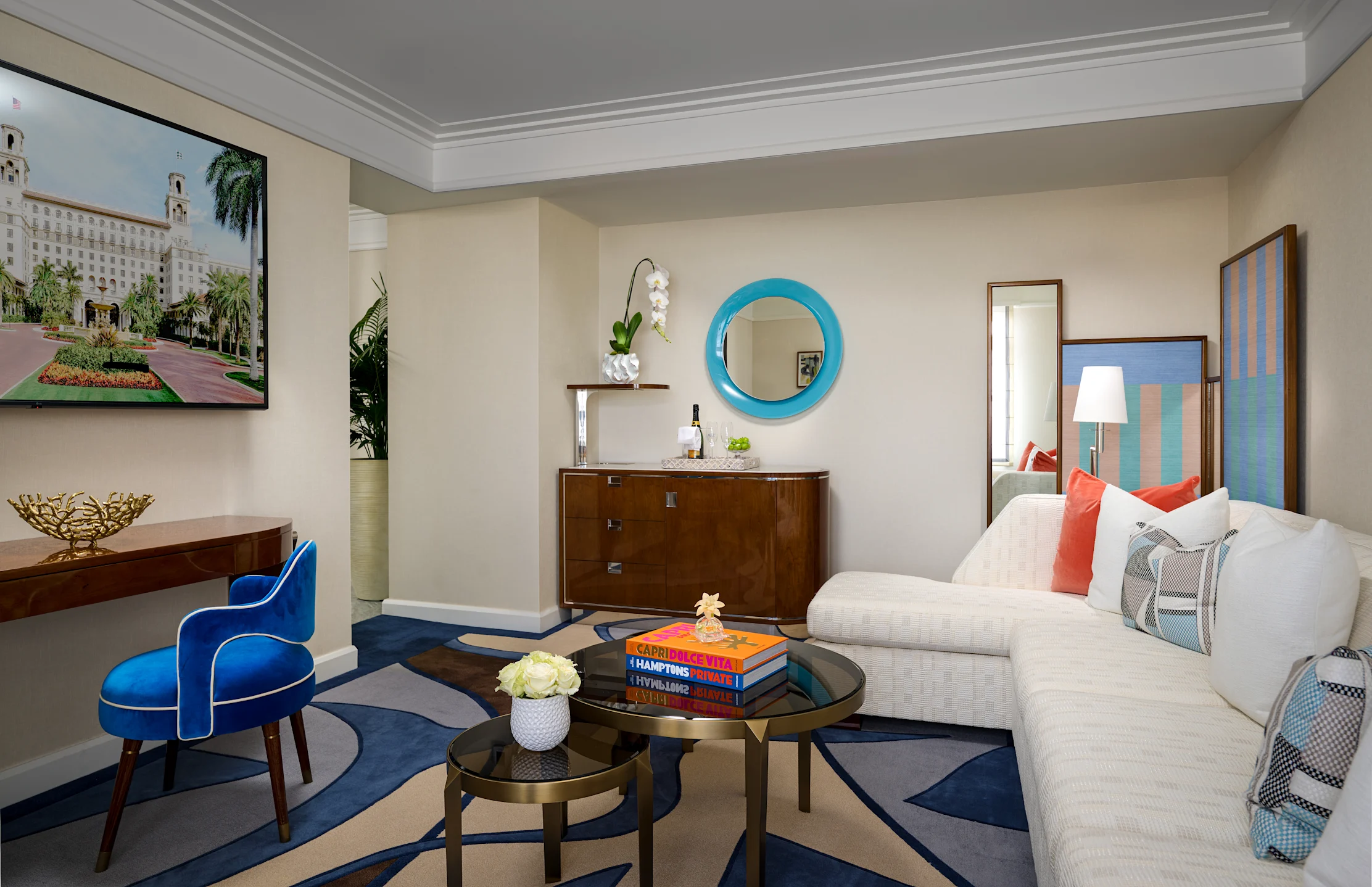 Modern hotel suite with beige walls, blue round mirror, white sofa, blue velvet chair, and navy rug.