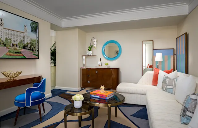 Modern hotel suite with beige walls, blue round mirror, white sofa, blue velvet chair, and navy rug.
