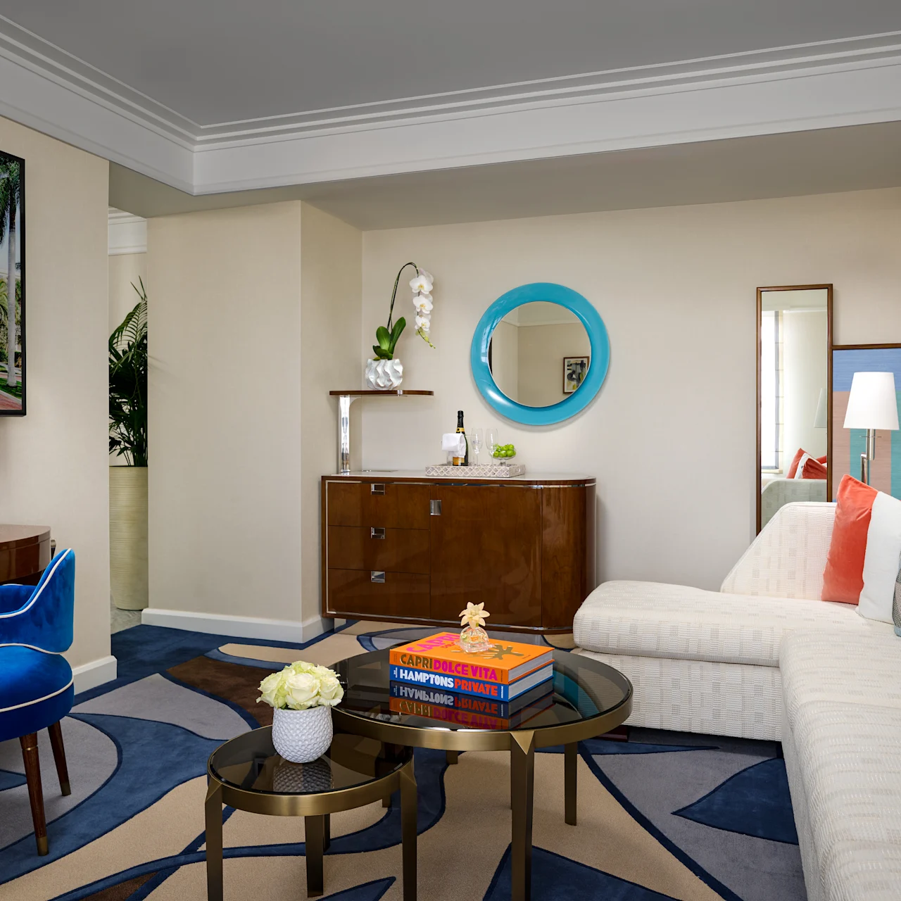 Modern hotel suite with beige walls, blue round mirror, white sofa, blue velvet chair, and navy rug.