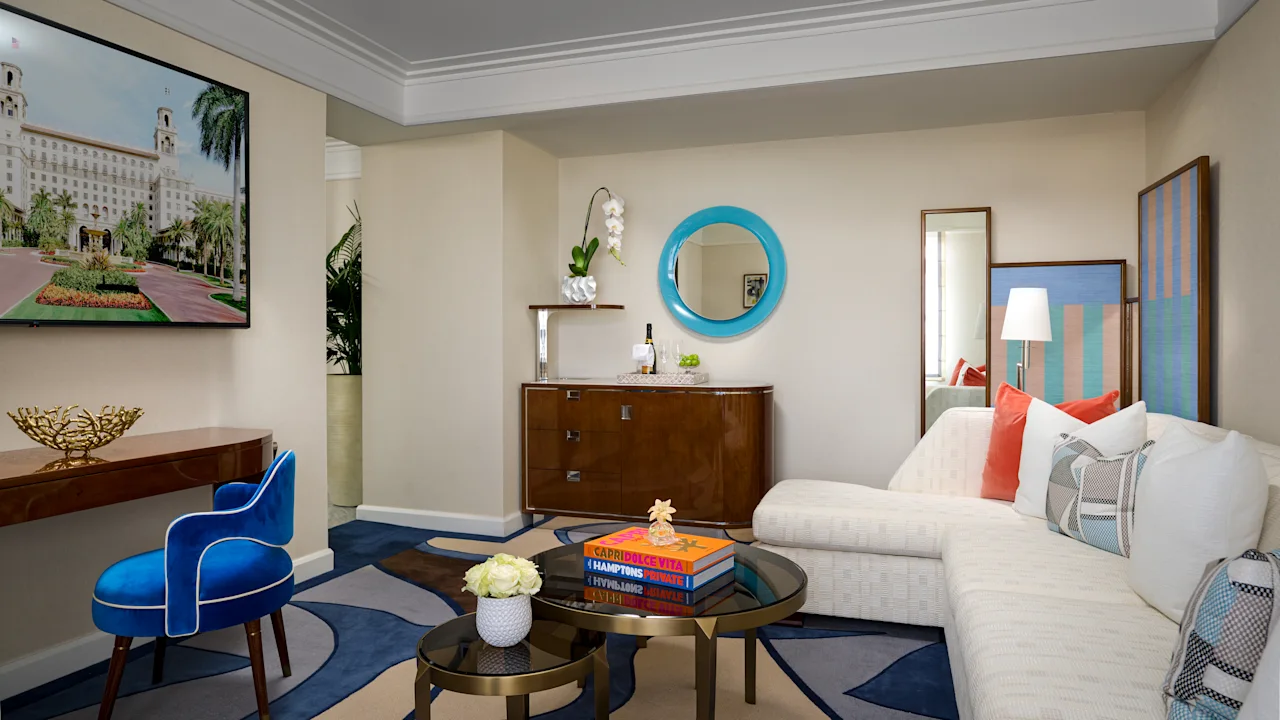 Modern hotel suite with beige walls, blue round mirror, white sofa, blue velvet chair, and navy rug.