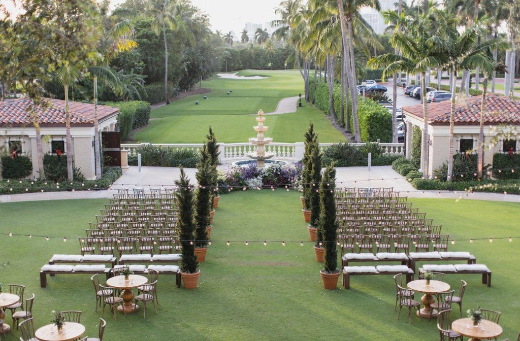 Outdoor wedding ceremony setup on green lawn with rows of chairs, fountain, palm trees, and buildings.