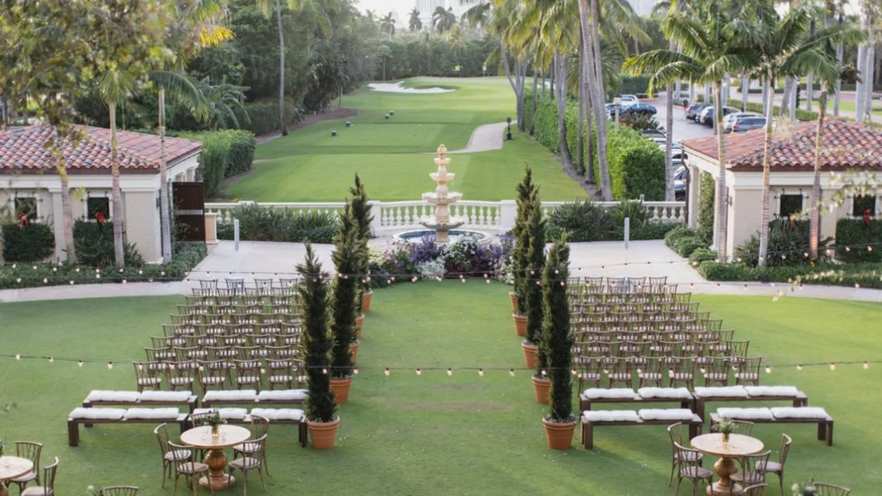 Outdoor wedding ceremony setup on green lawn with rows of chairs, fountain, palm trees, and buildings.