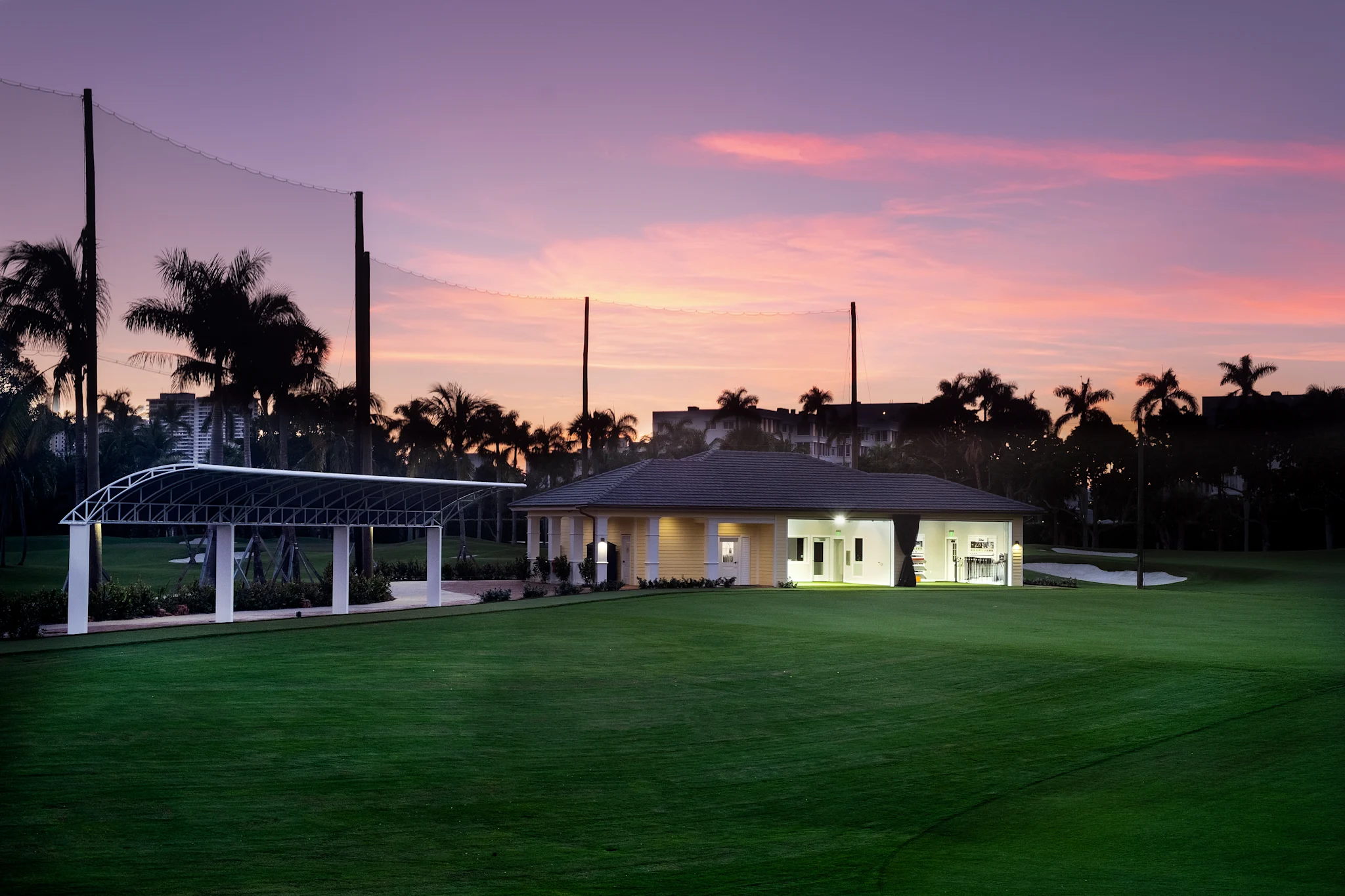 Illuminated clubhouse and pavilion on green lawn at dusk with pink and purple sky and palm trees.