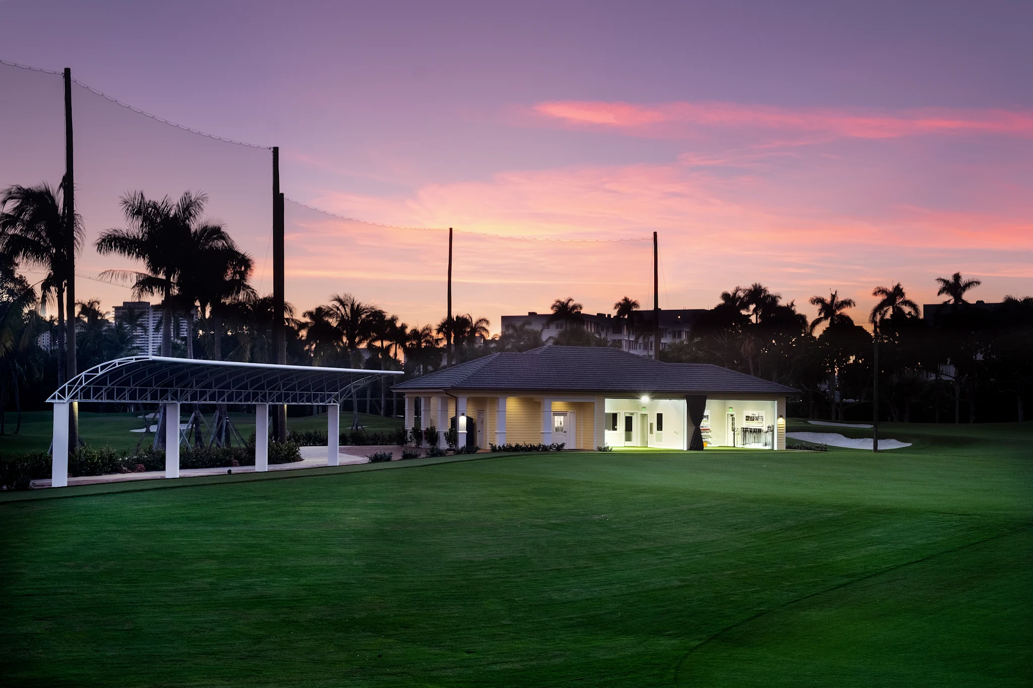 Illuminated clubhouse and pavilion on green lawn at dusk with pink and purple sky and palm trees.