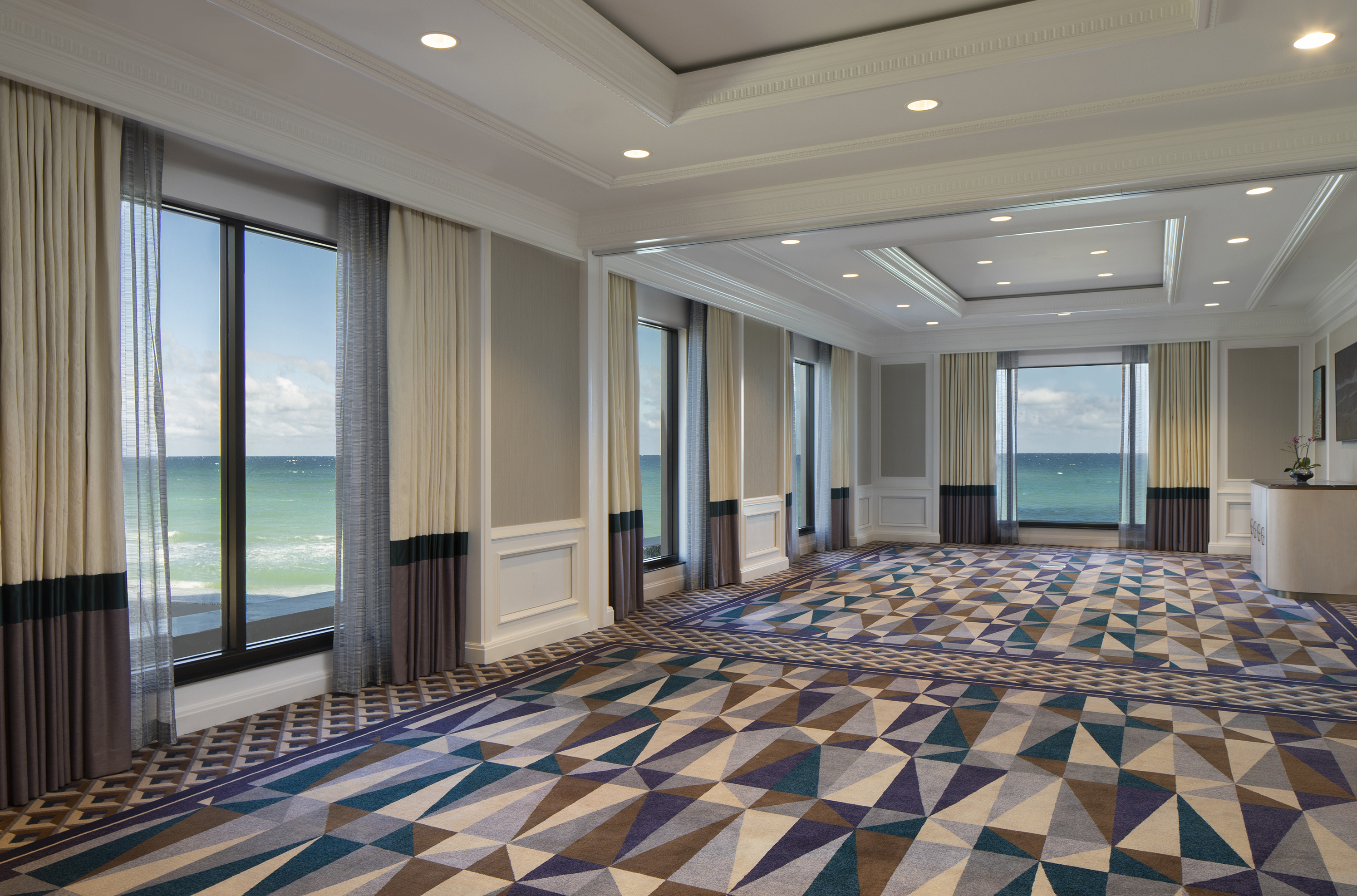 Elegant ballroom with geometric patterned carpet, floor-to-ceiling windows, and ocean views.