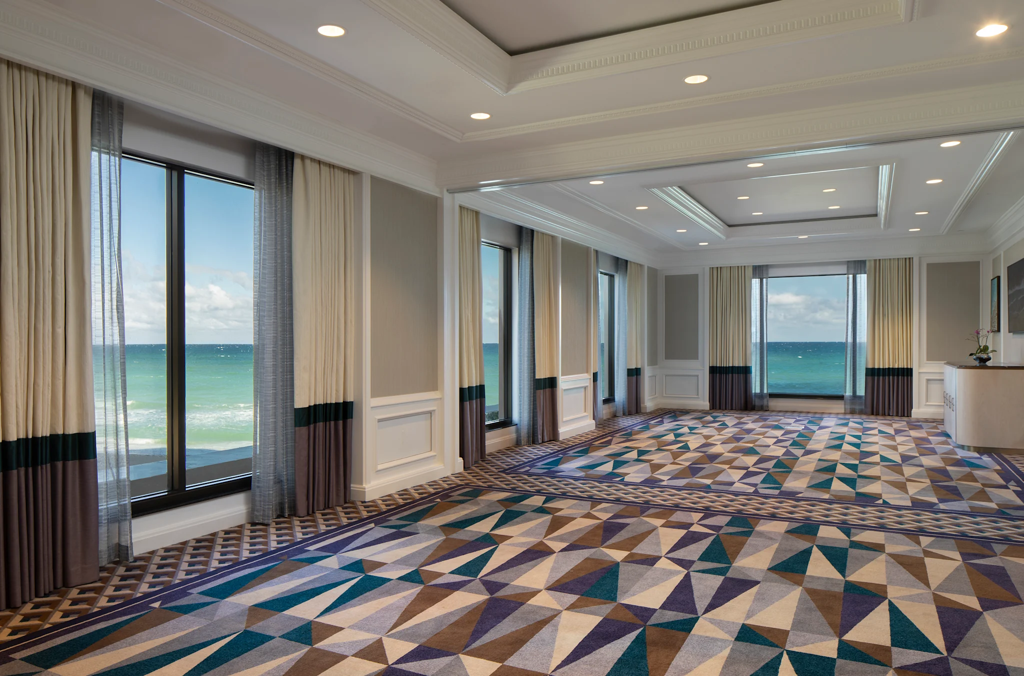 Elegant ballroom with geometric patterned carpet, floor-to-ceiling windows, and ocean views.