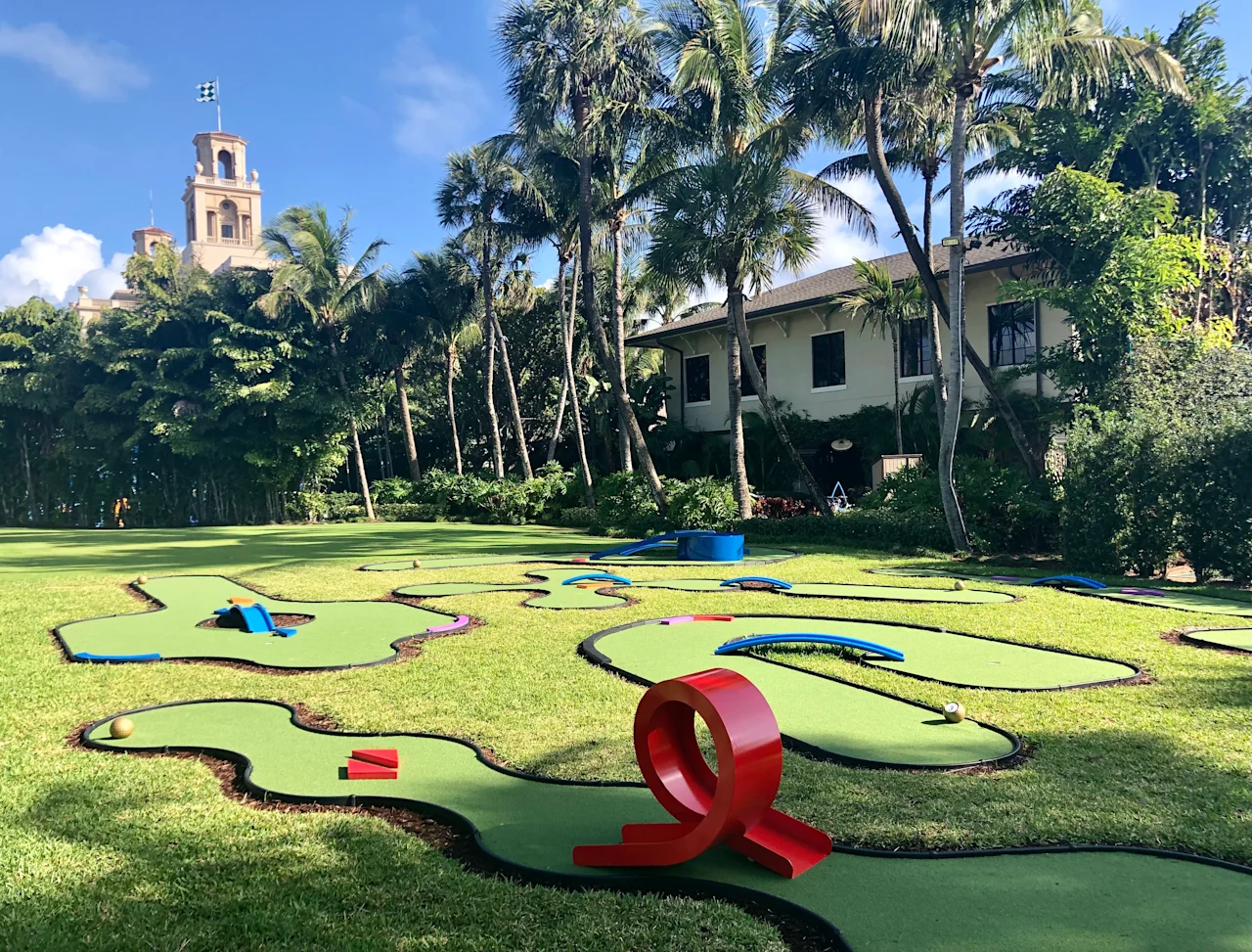 Miniature golf course with colorful obstacles on green lawn, palm trees and bell tower in background.