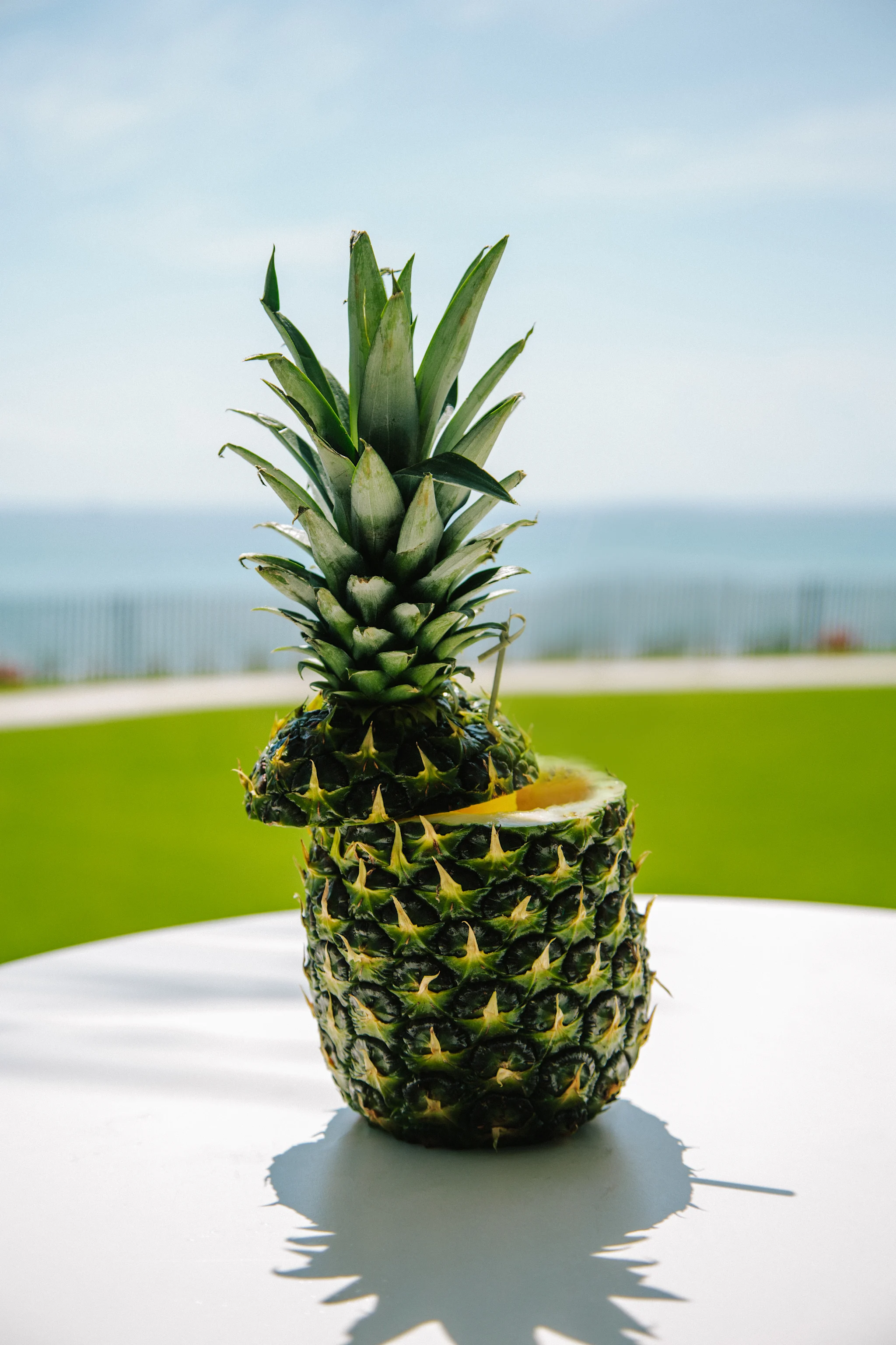 Pineapple with a slice cut out on a white table with ocean and green lawn in the background.