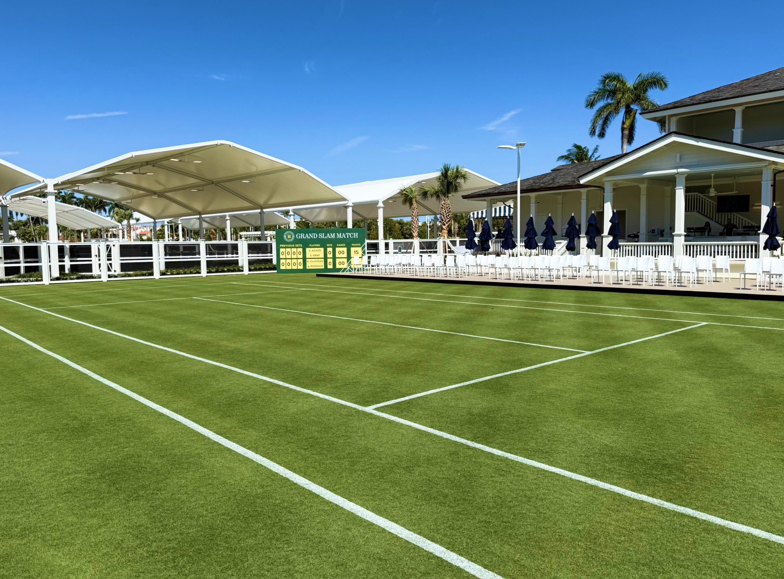 Grass tennis court with white canopy, scoreboard, and clubhouse with palm trees under blue sky.