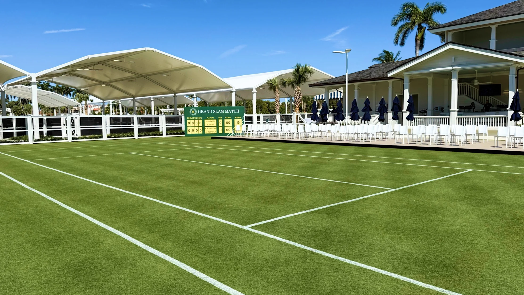 Grass tennis court with white canopy, scoreboard, and clubhouse with palm trees under blue sky.