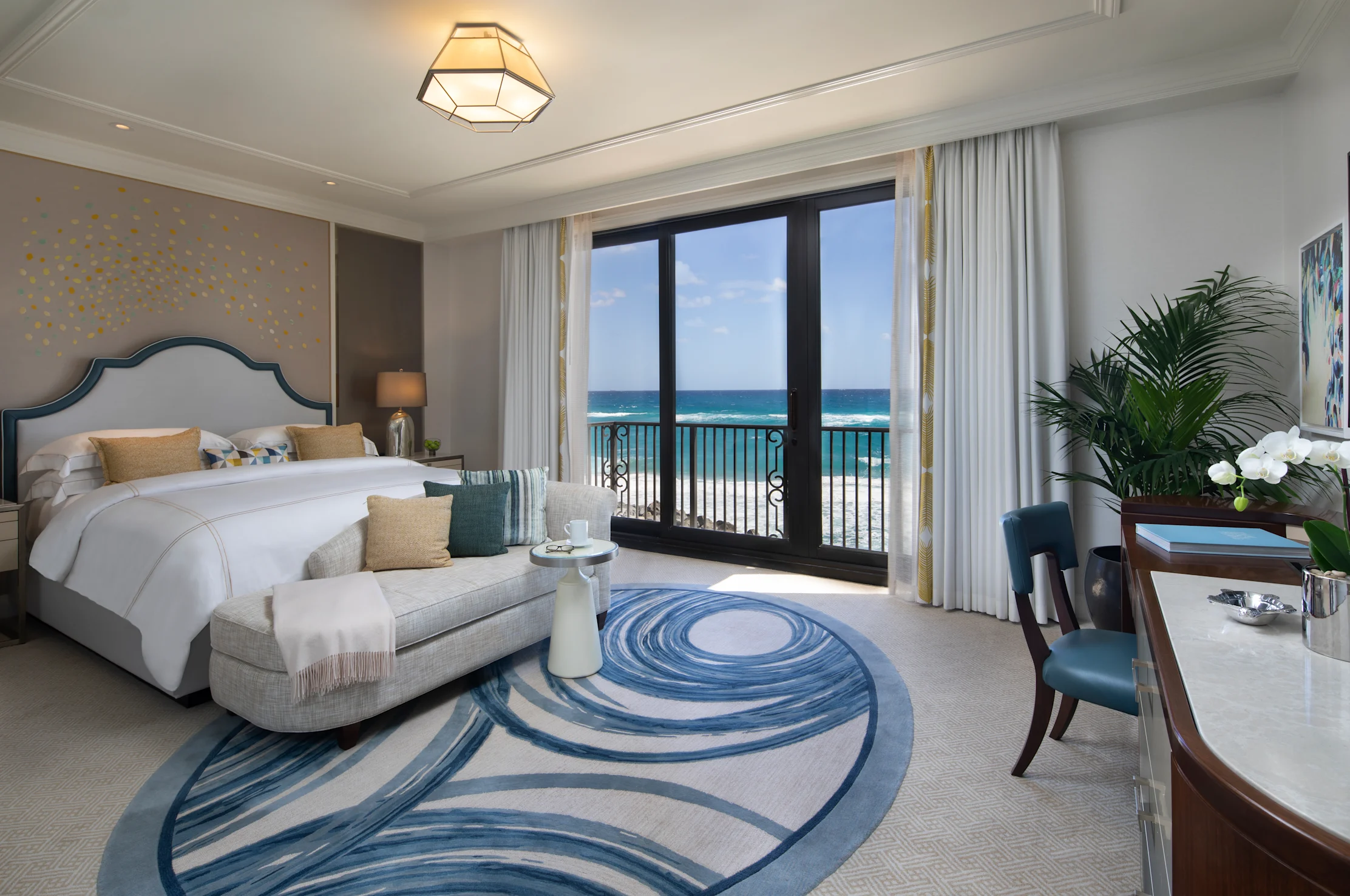 Luxury hotel bedroom with ocean view through balcony doors, featuring white bedding and blue accent rug.