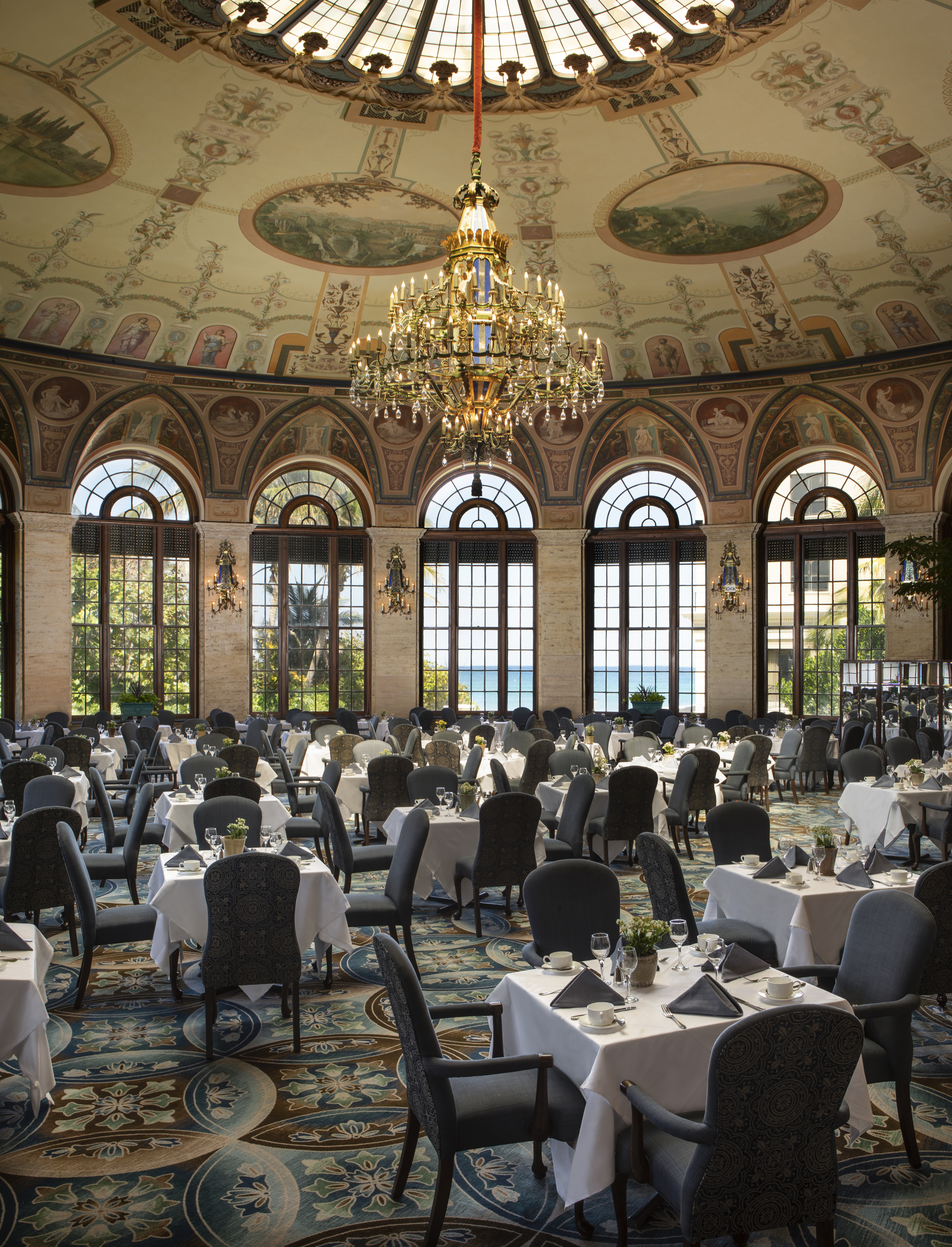 Ornate dining room with painted dome ceiling, grand chandelier, arched windows, and formal table settings.