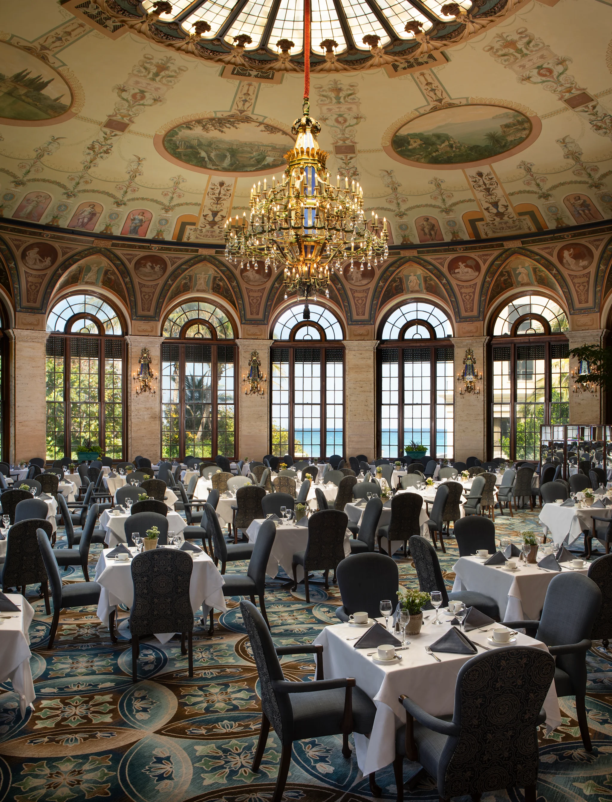 Ornate dining room with painted dome ceiling, grand chandelier, arched windows, and formal table settings.