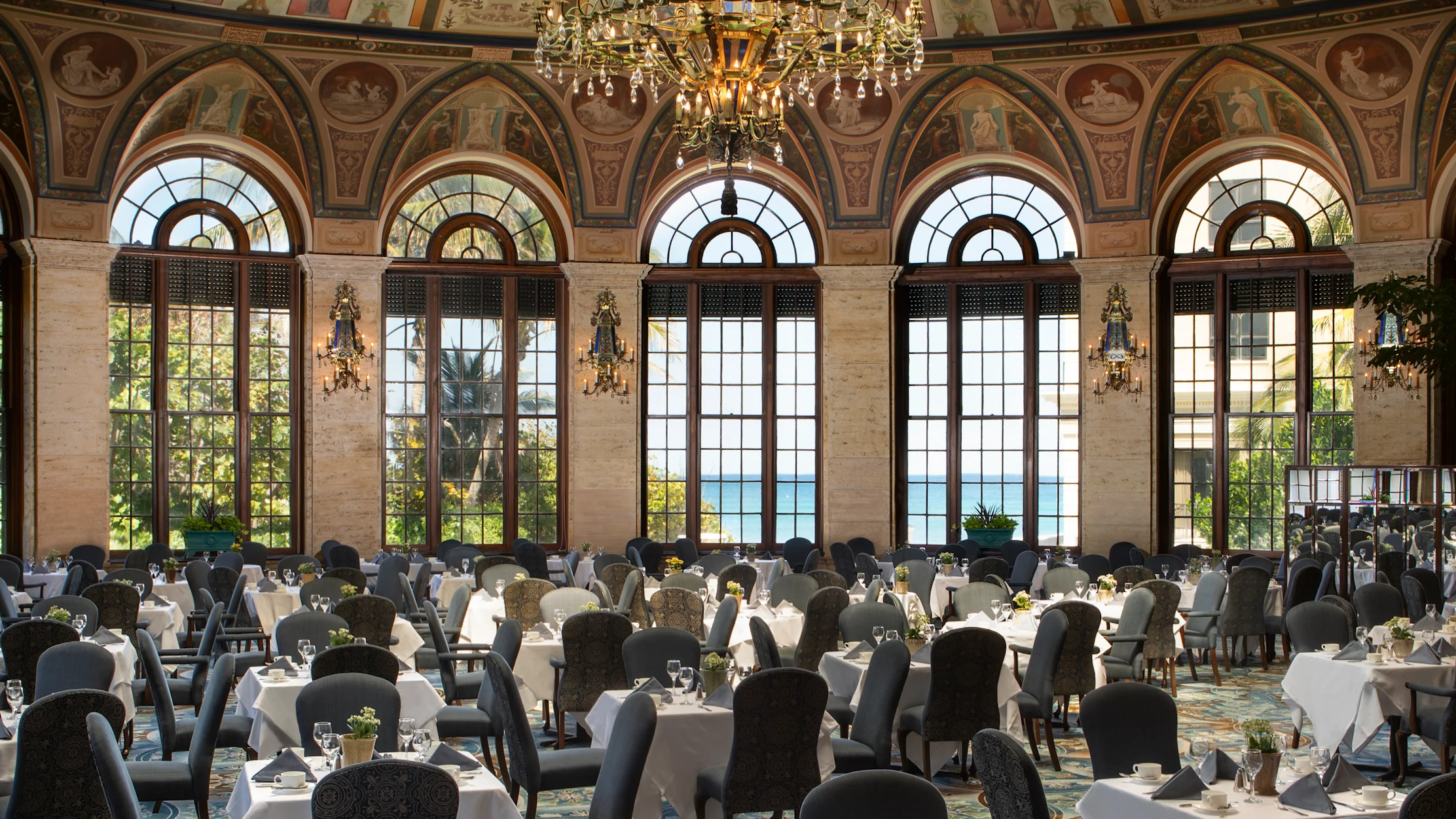Ornate dining room with painted dome ceiling, grand chandelier, arched windows, and formal table settings.