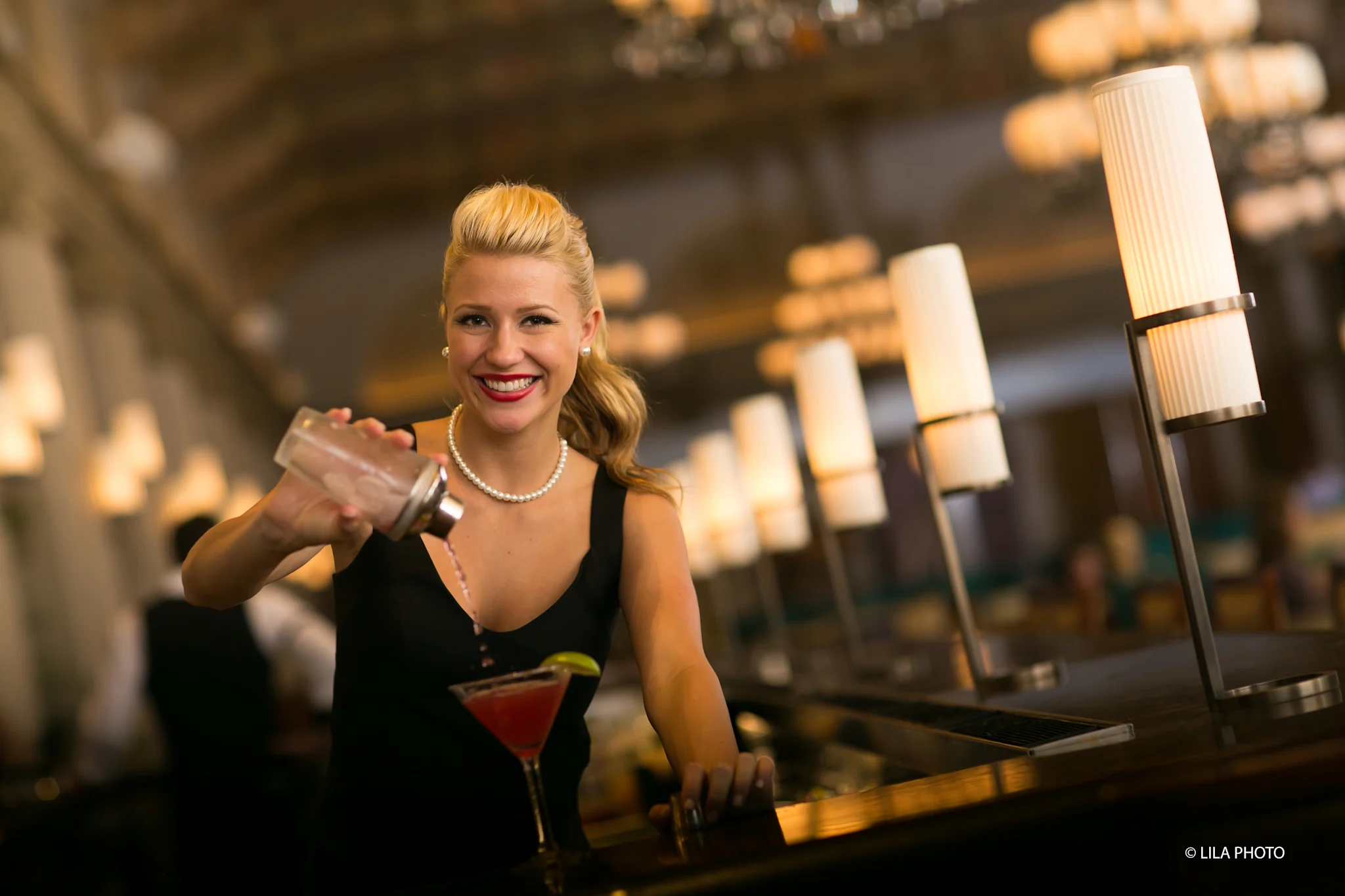 Smiling blonde bartender in black dress and pearls shaking a cocktail in an upscale bar setting.