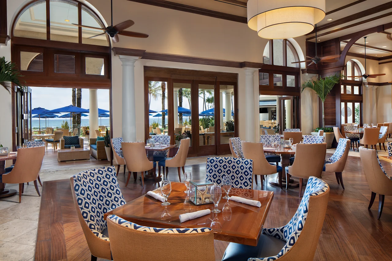 Upscale restaurant interior with tan and blue patterned chairs, wooden tables, arched windows, and ocean view.