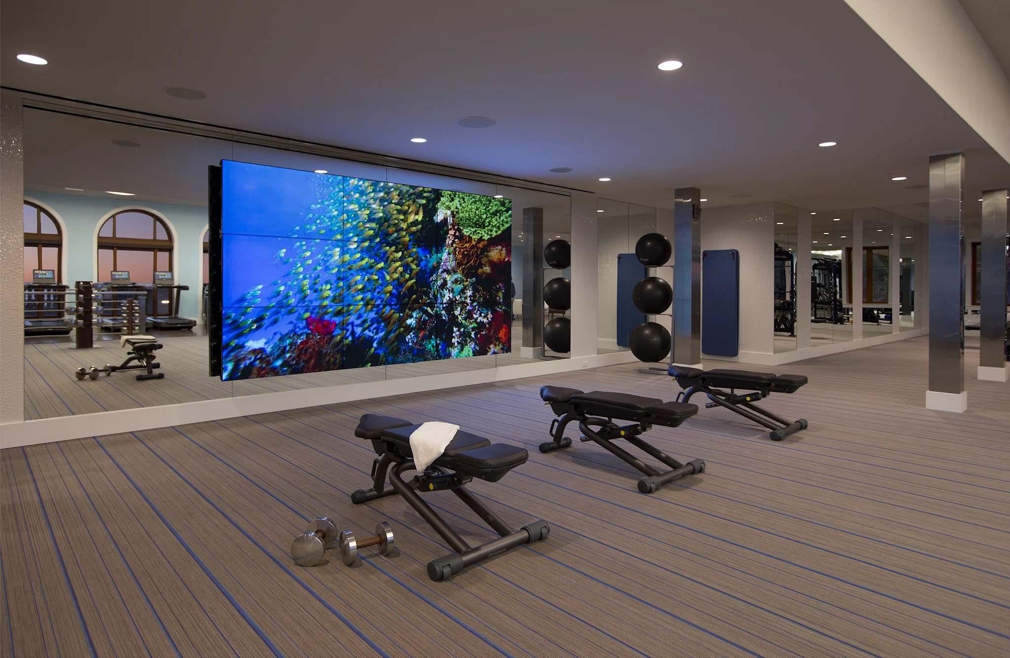 Modern gym with exercise benches, weights, and large screen displaying colorful underwater coral reef.